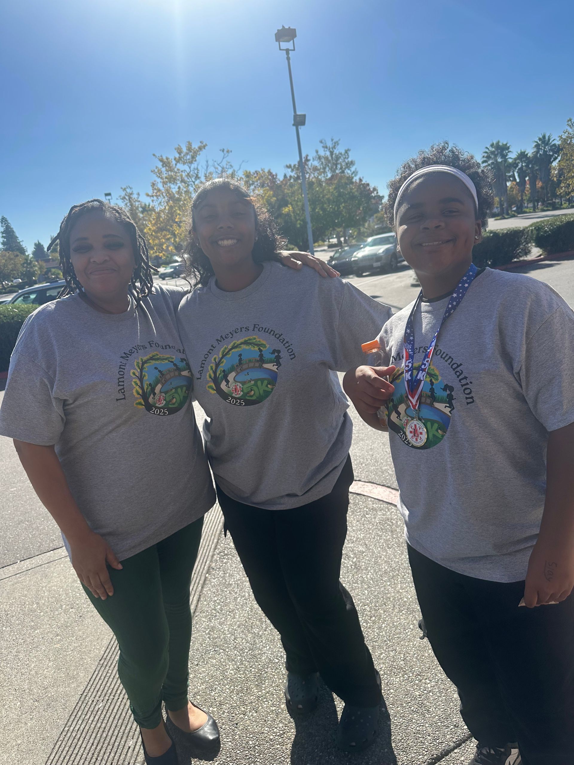 Three people in gray shirts pose outdoors, smiling. They stand under a blue sky, near a parking lot.