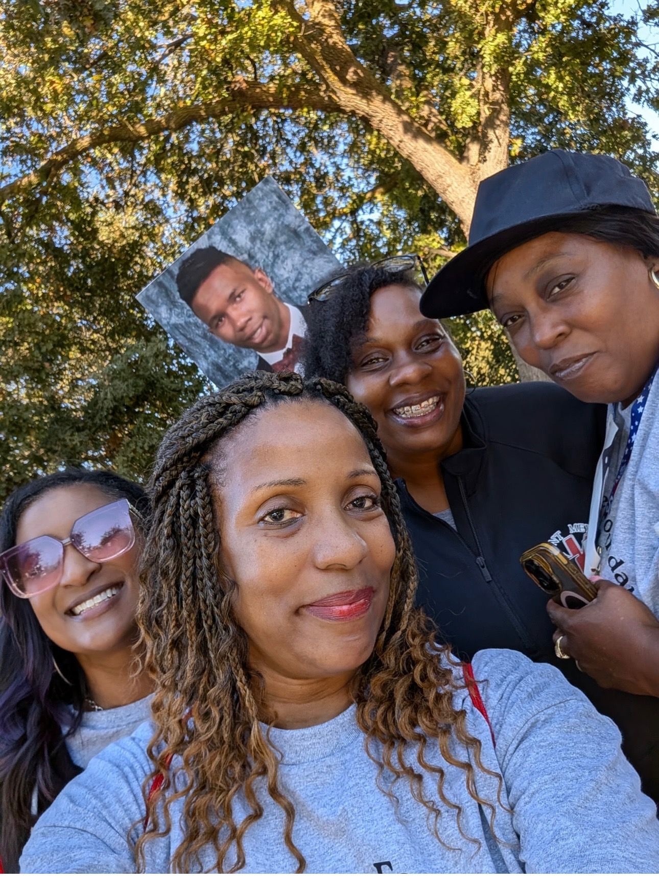 Four women smile, holding a photo of a young man. Outdoors, sunny.