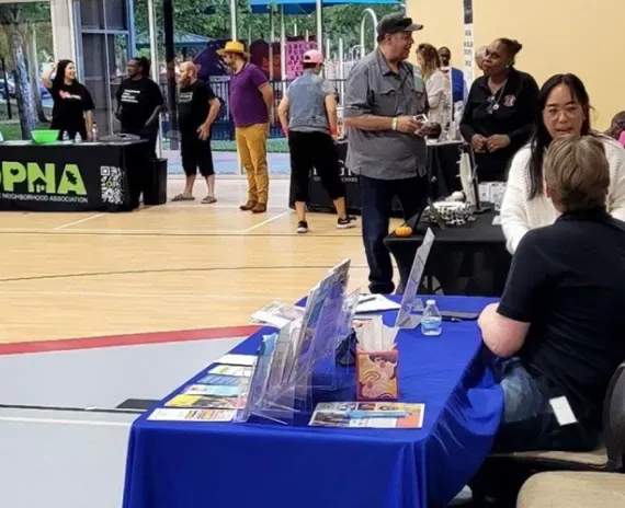 People at booths in a gym, attending an event. Tables with pamphlets; PNA logo visible.