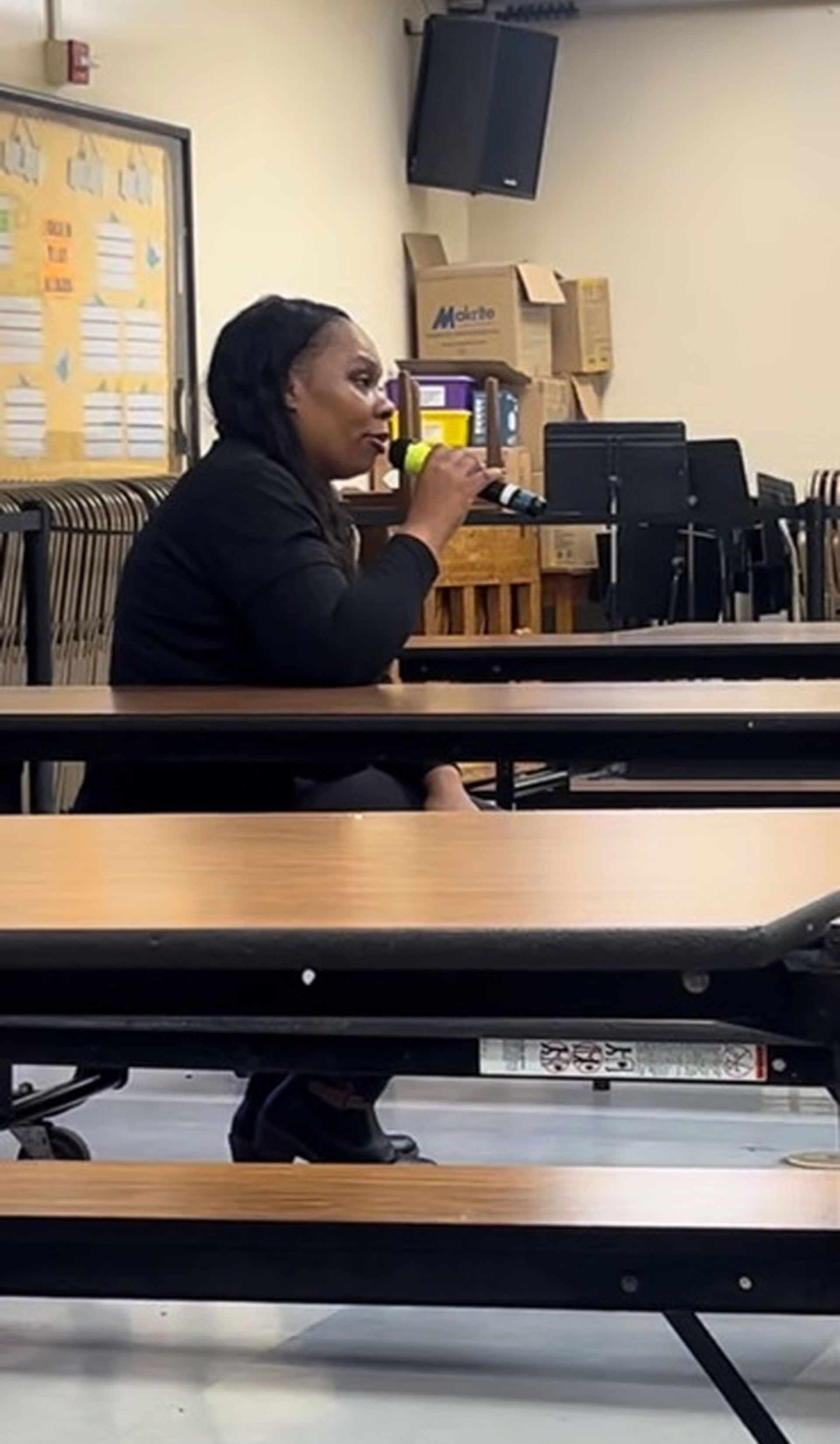 A woman is sitting at a table in a classroom drinking from a cup.