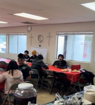 People seated at tables with red tablecloths, inside a room, possibly eating. Windows and a clock are visible.