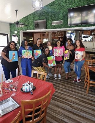 Group of people at a paint and sip event, holding up their artwork. Red table, paintings, and green wall in the background.