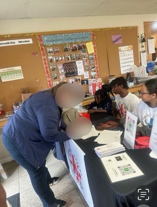 People at a table with promotional materials. One person inspects a helmet, others sit. Bulletin board in background.