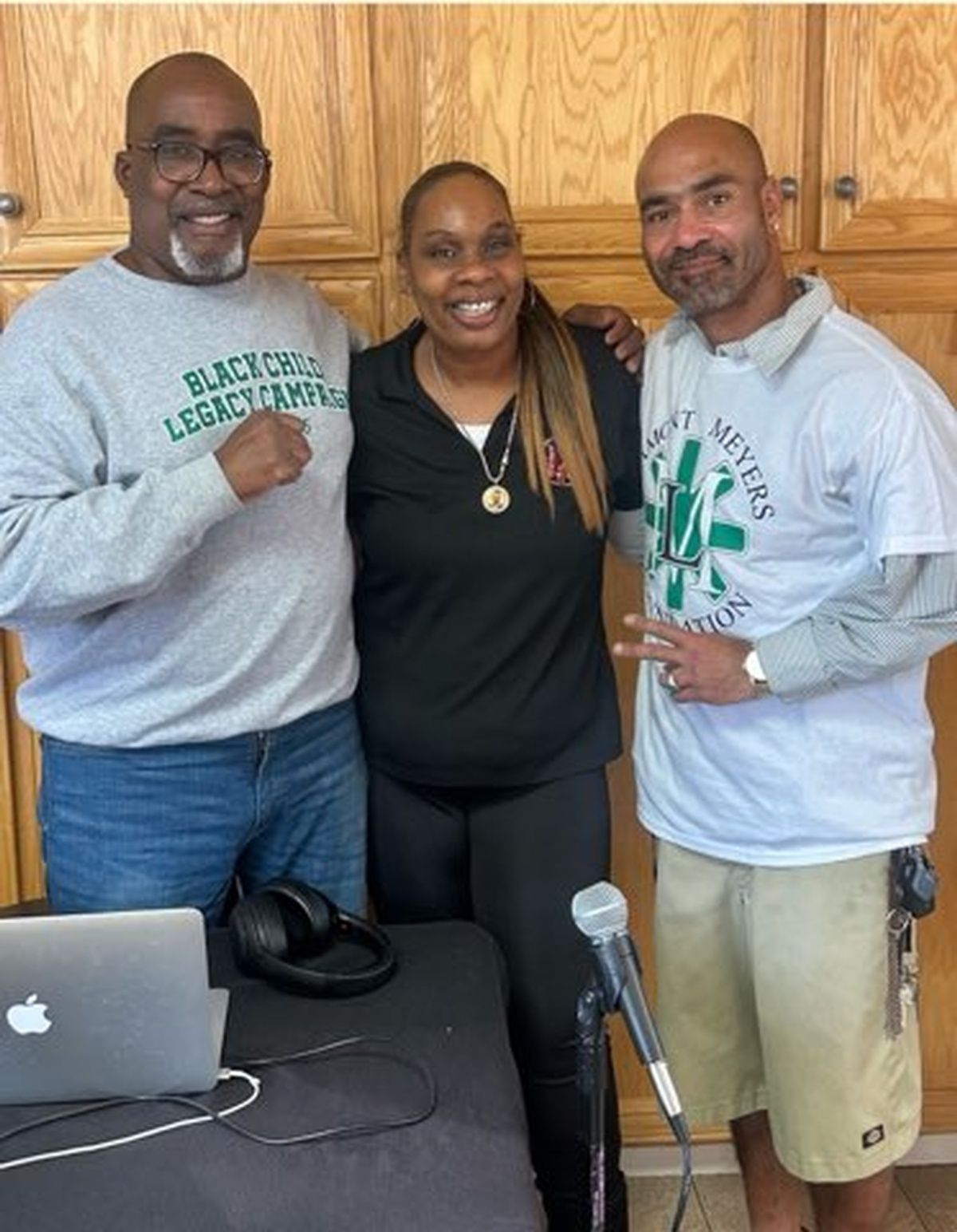 Three people posing by a table with a laptop and microphone. They are smiling; one person is wearing a shir