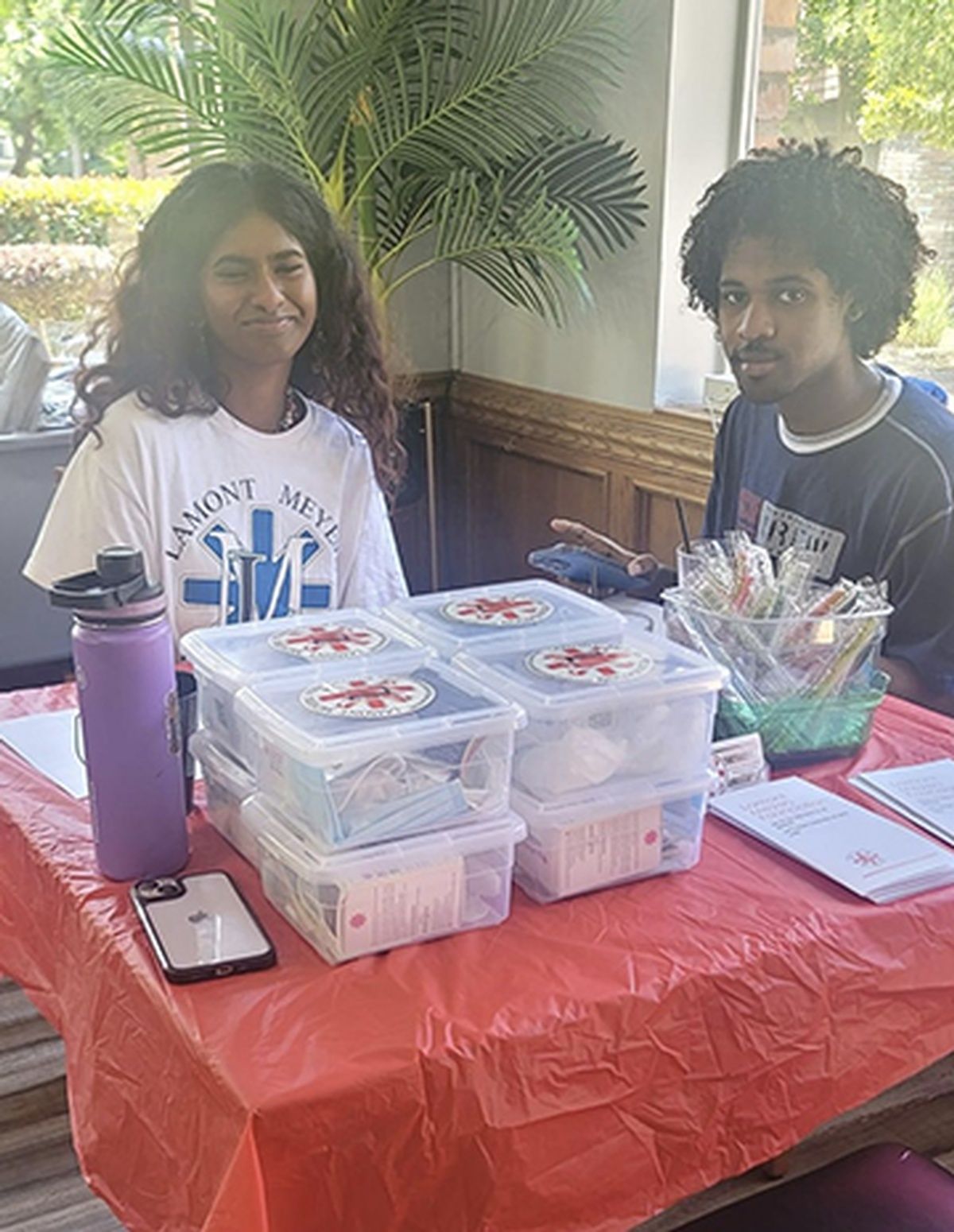 Two people at a table with medical supplies. They are smiling, with a red tablecloth and a building in the background.