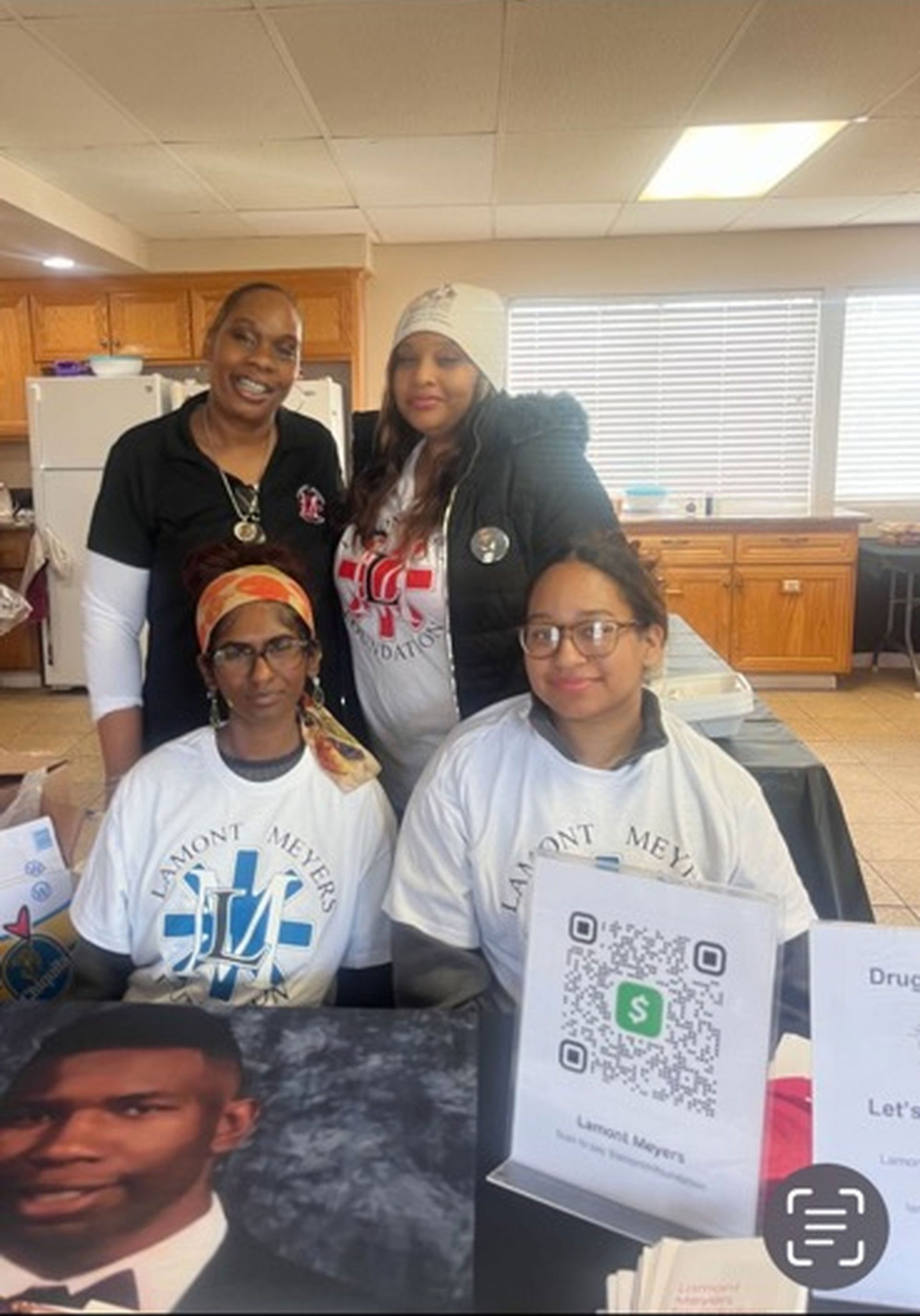 Four people pose near a table with a portrait and QR code. Indoors, they wear shirts with a logo.