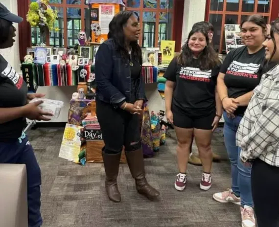 Woman in a denim jacket speaks to a group of students in a library.
