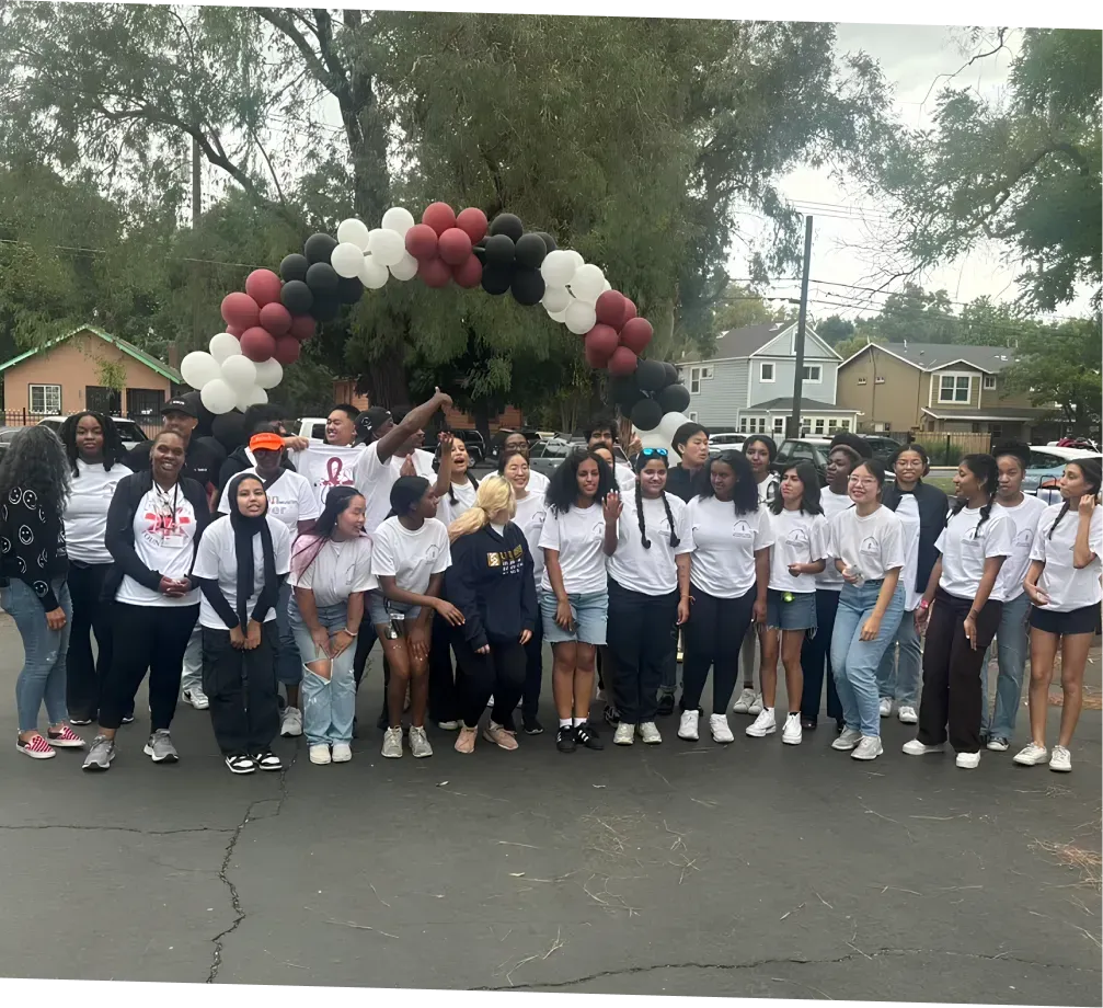 Group of people under a balloon arch, some smiling, in front of houses on a street.