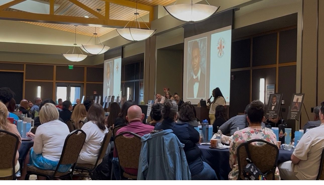 Audience at a conference listens to a presentation on a screen. Large room with a stage, ceiling lights.
