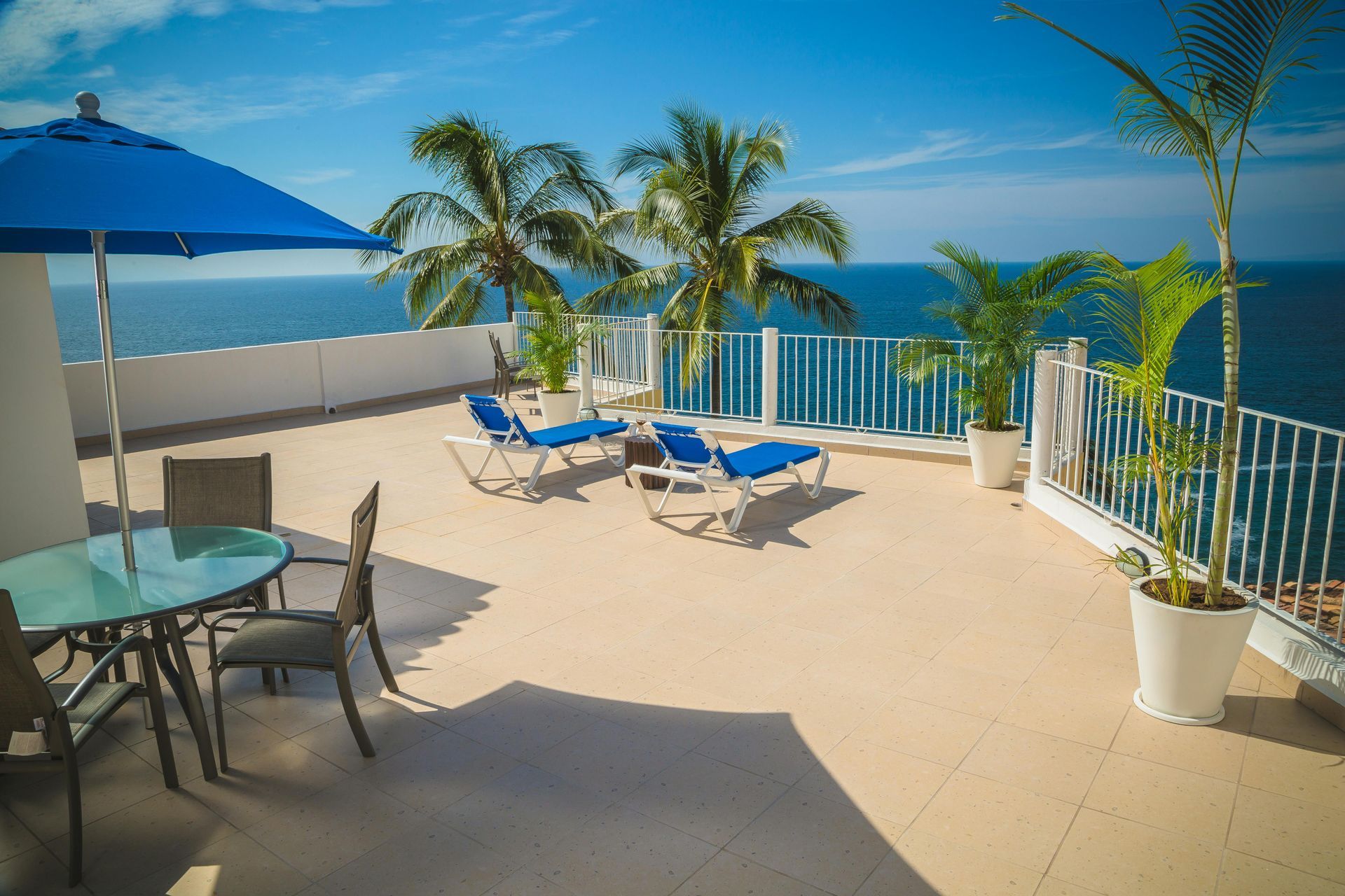 Rooftop terrace overlooking ocean; blue umbrella, lounge chairs, table, palm trees, white railing, sunny day.