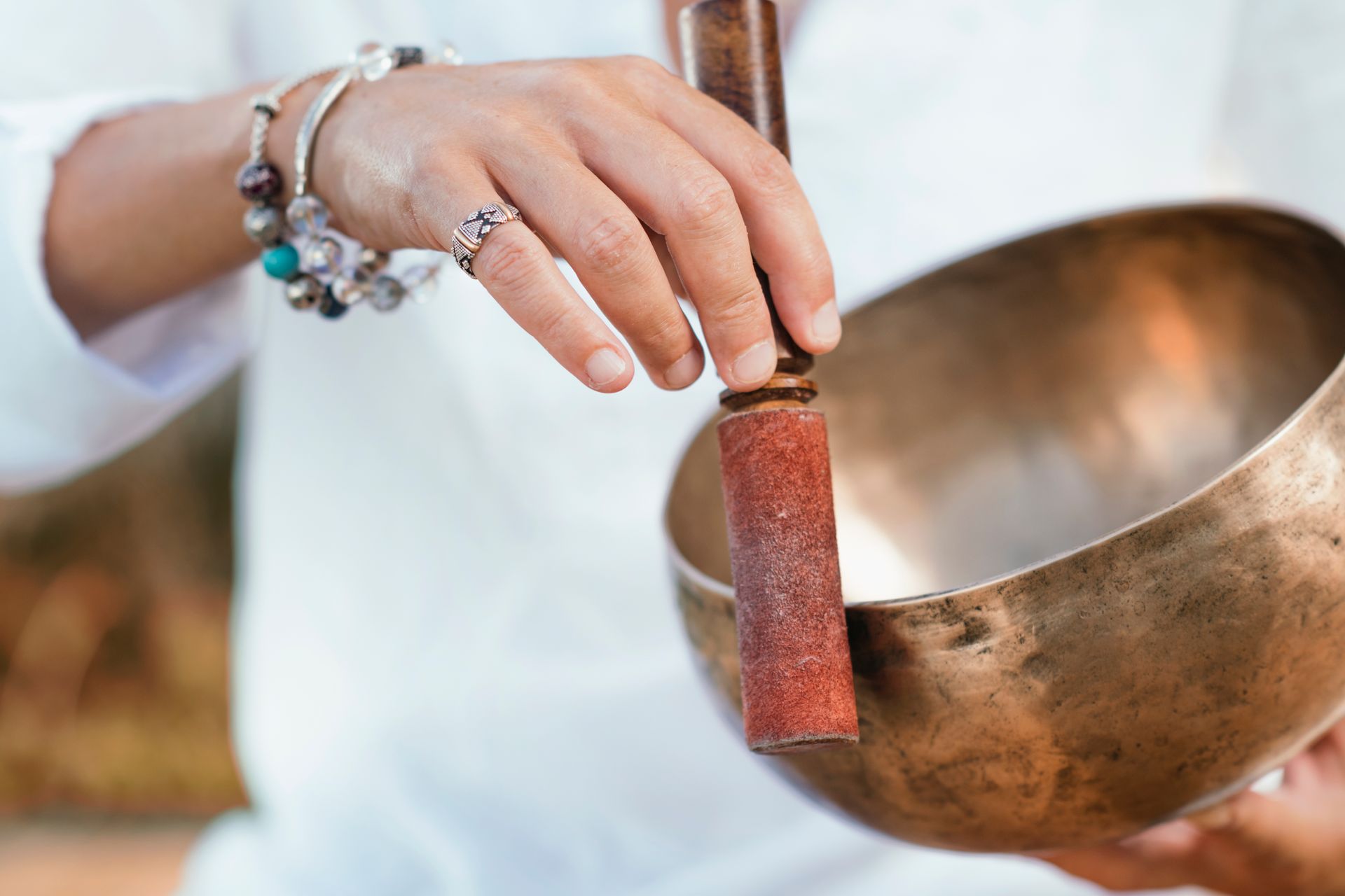 Woman holding a singing bowl, striking it with a mallet. She wears bracelets and a ring; outdoors.