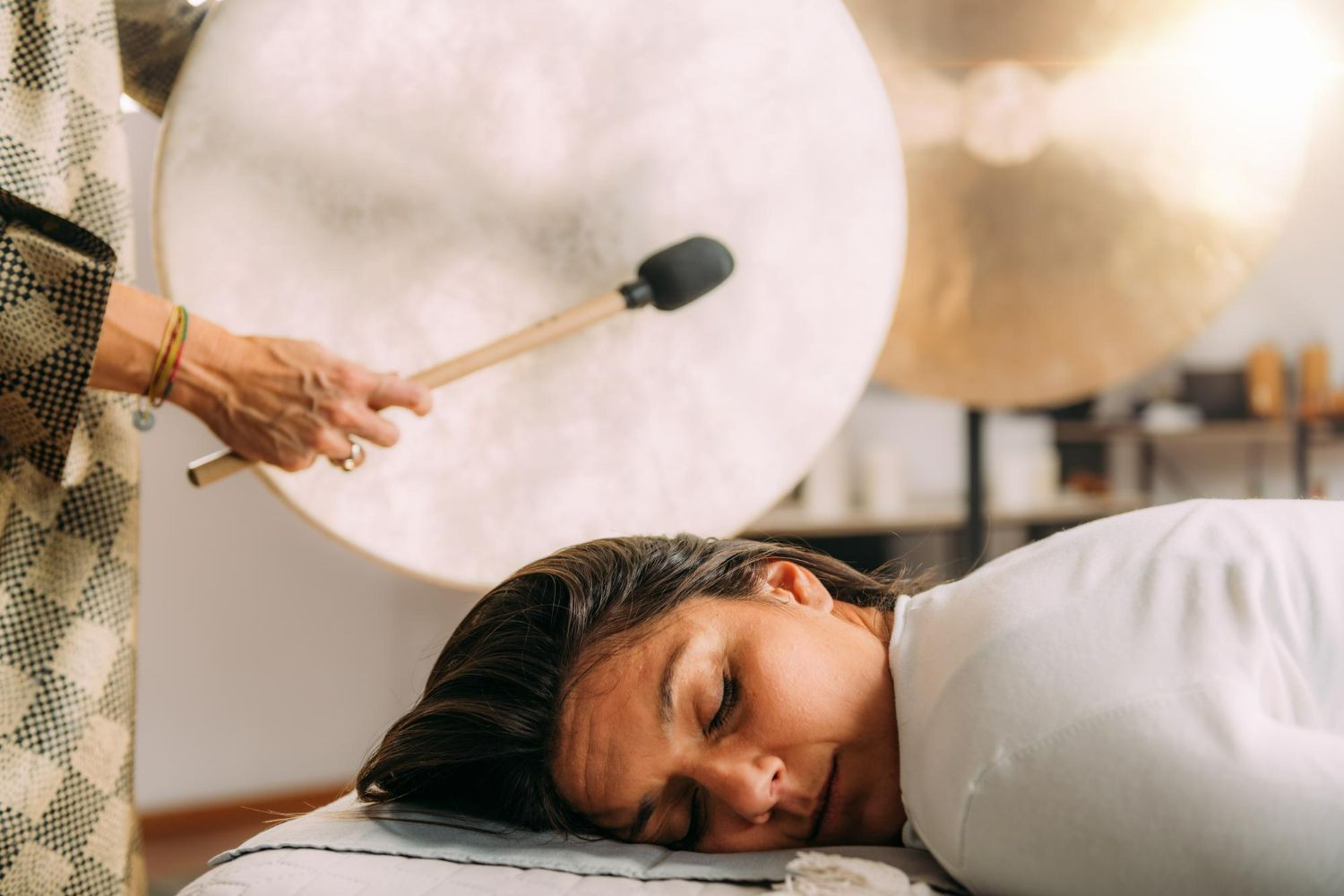 Person lies on table as hand strikes gong with mallet; in room.