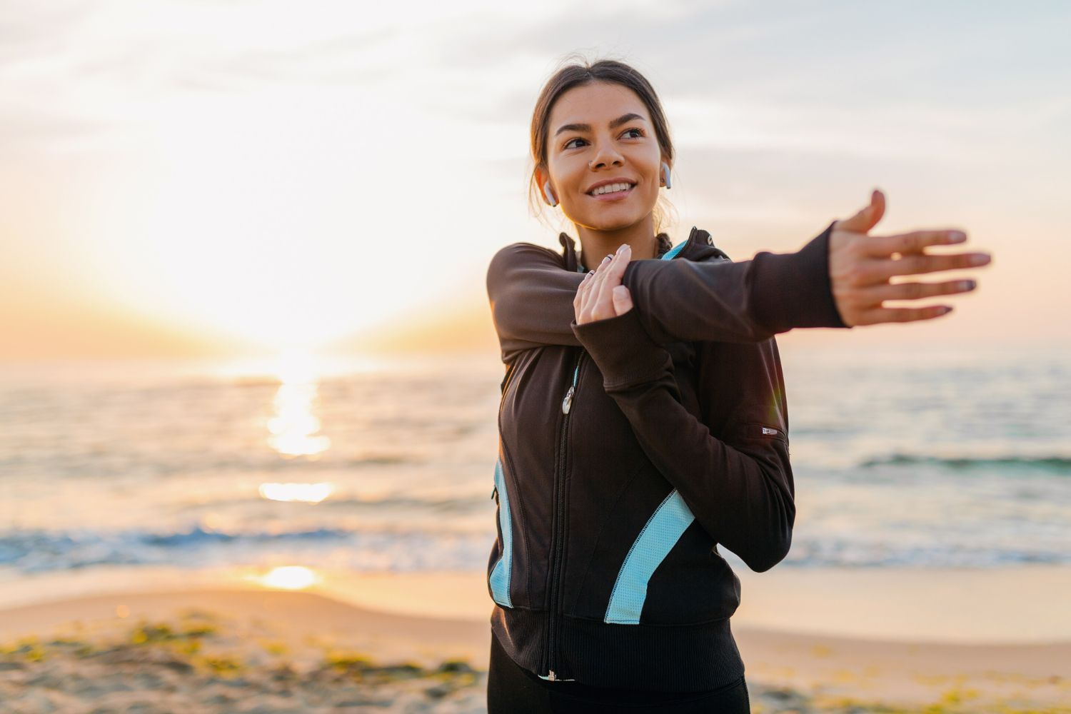 Woman stretching arm on beach at sunrise, wearing athletic jacket.