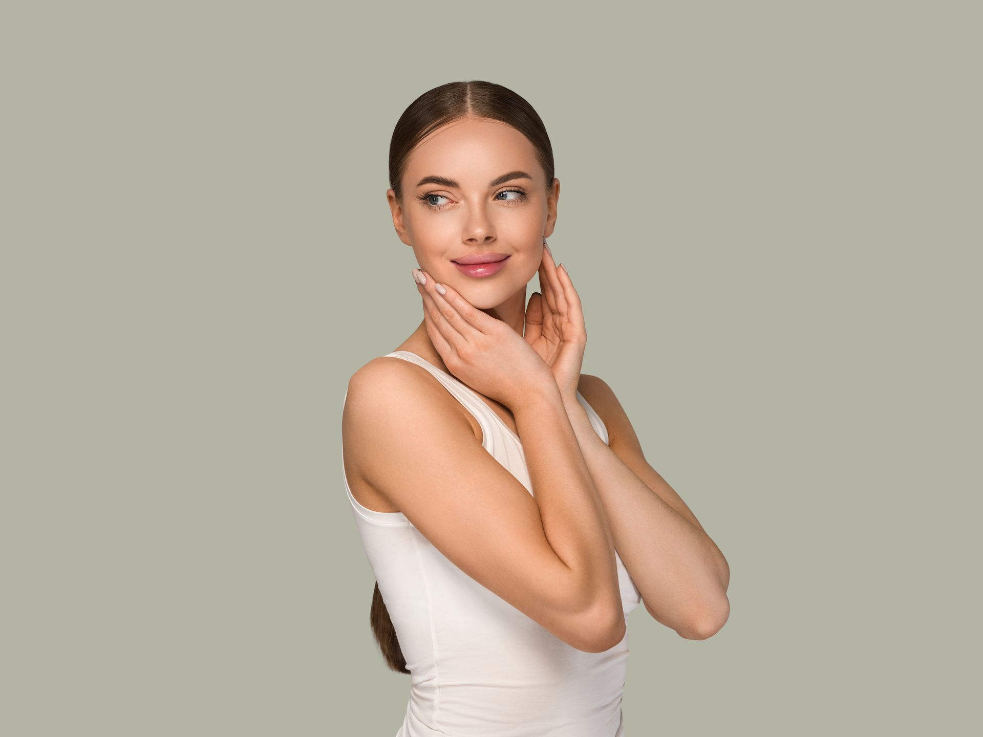 A woman in a white tank top is touching her face and smiling.