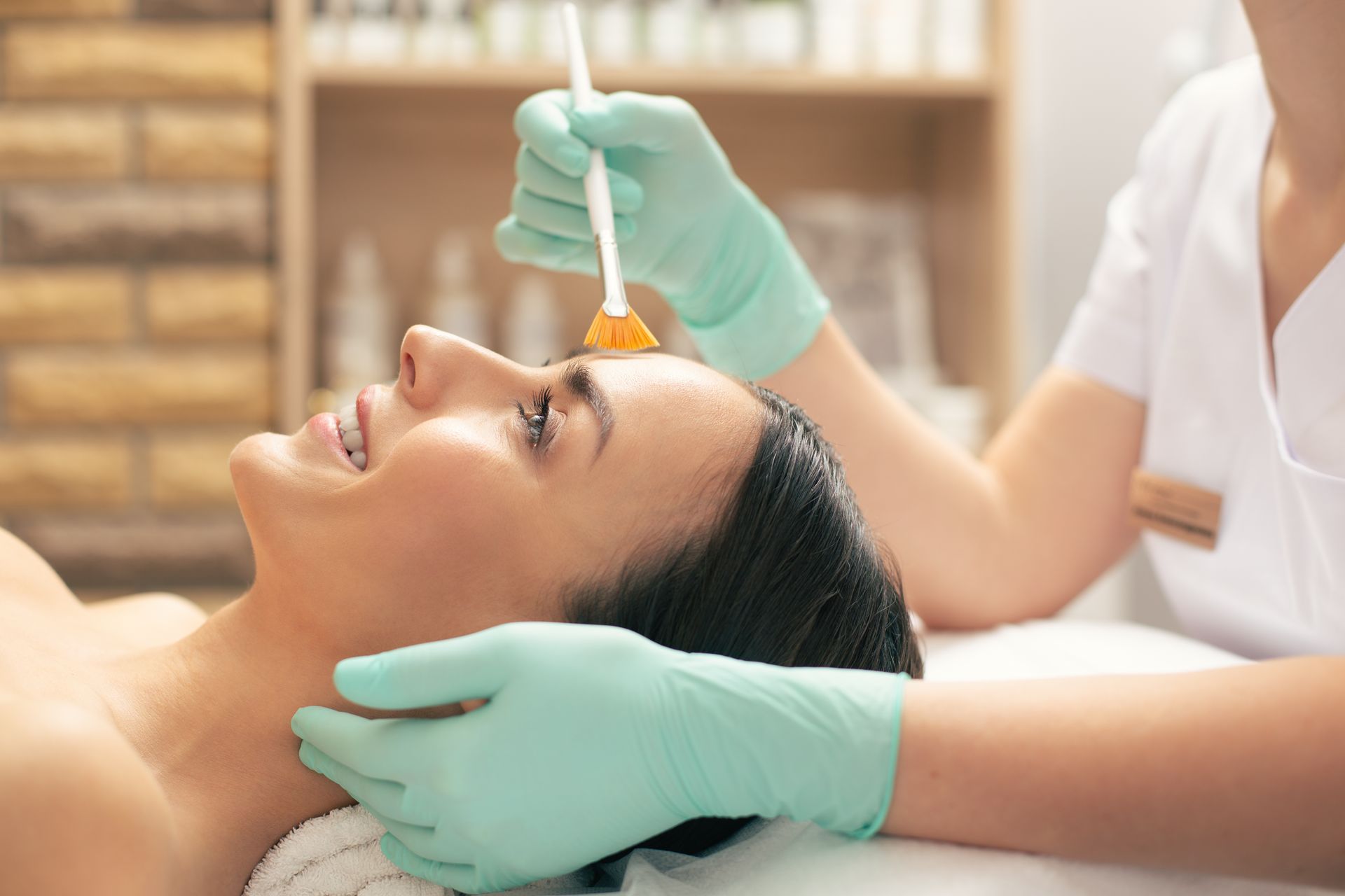 Woman receiving facial treatment, professional applying product to forehead with a brush, light green gloves.