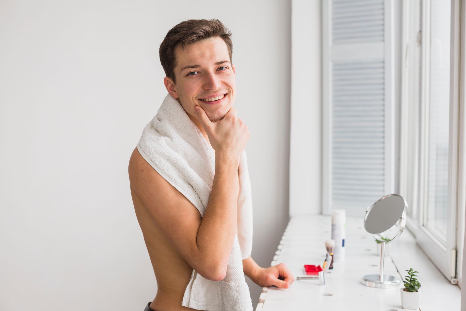 A shirtless man with a towel around his neck is looking at himself in the mirror.