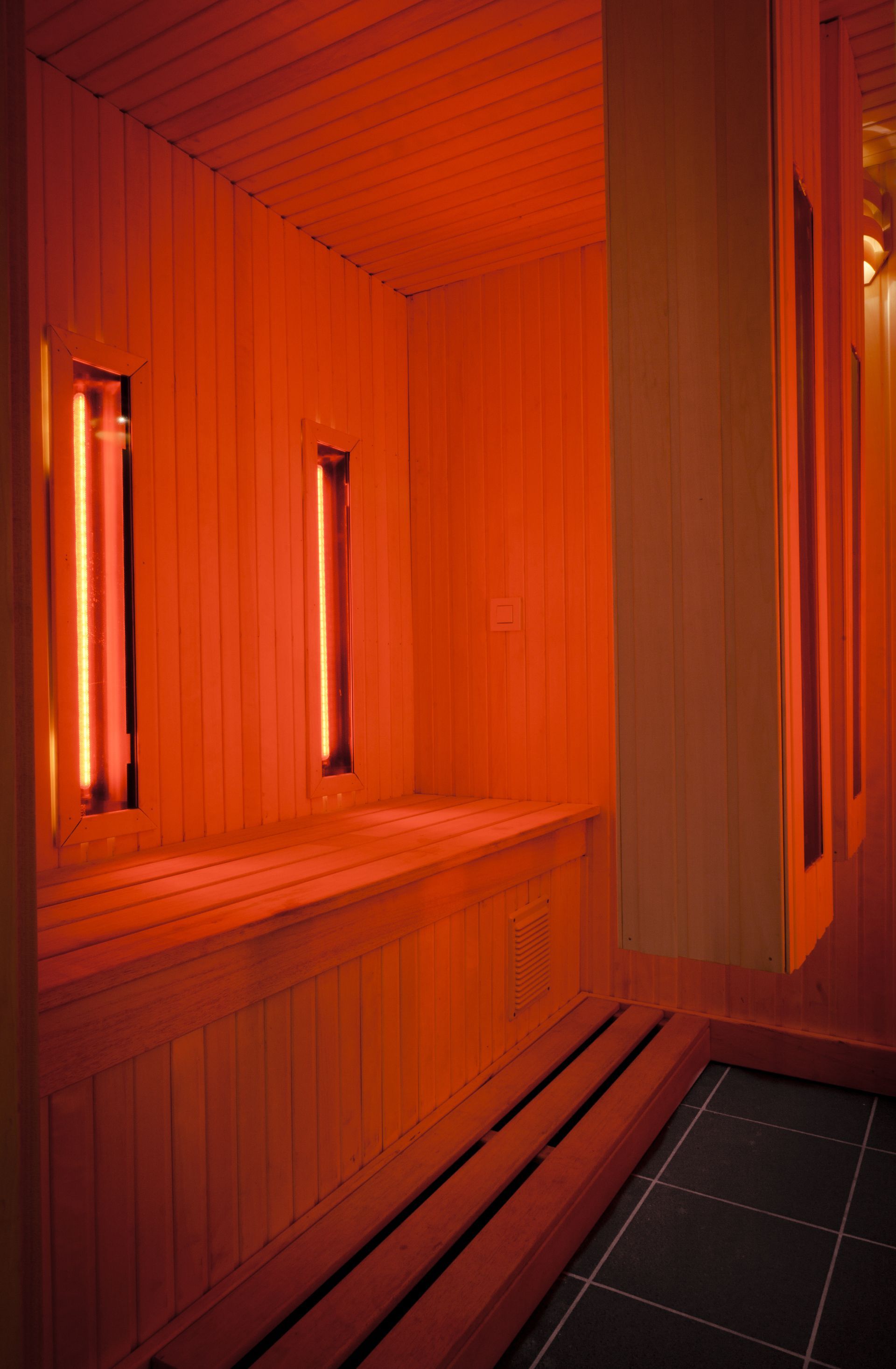 Inside a red-lit sauna; wooden benches, vertical heaters, and a black tiled floor.