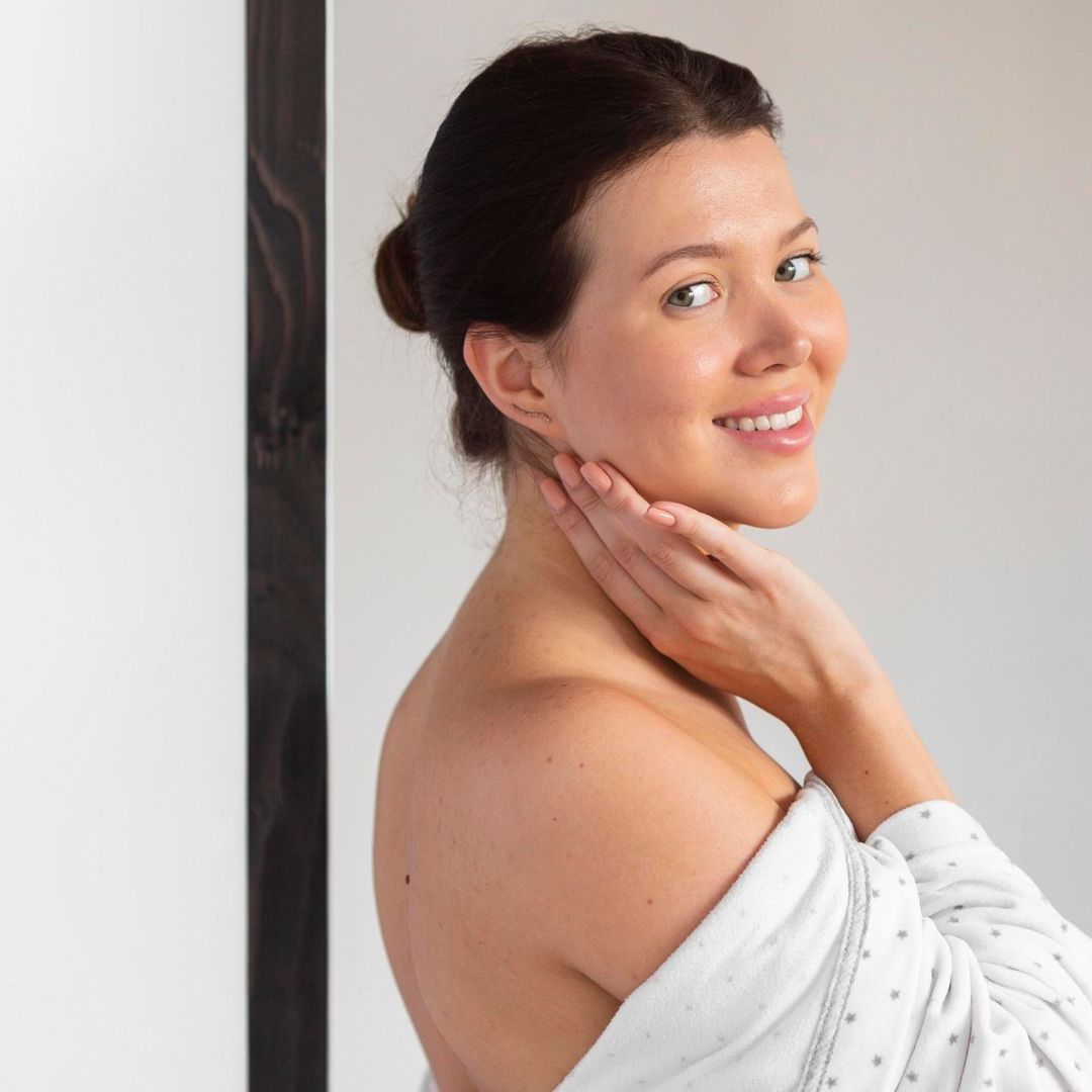 A woman in a white towel is smiling and touching her neck