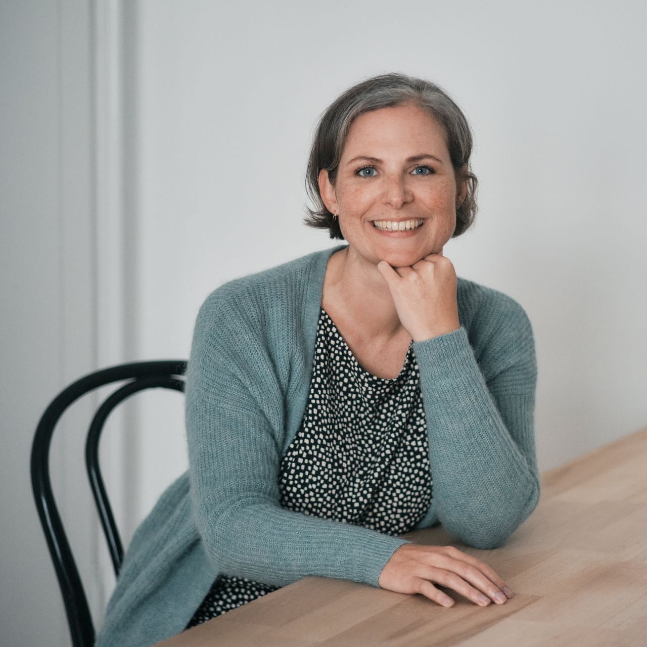 Frau mit kurzen grauen Haaren lächelt und lehnt an einem Tisch. Trägt eine blaue Strickjacke und ein gemustertes Oberteil. Drinnen.