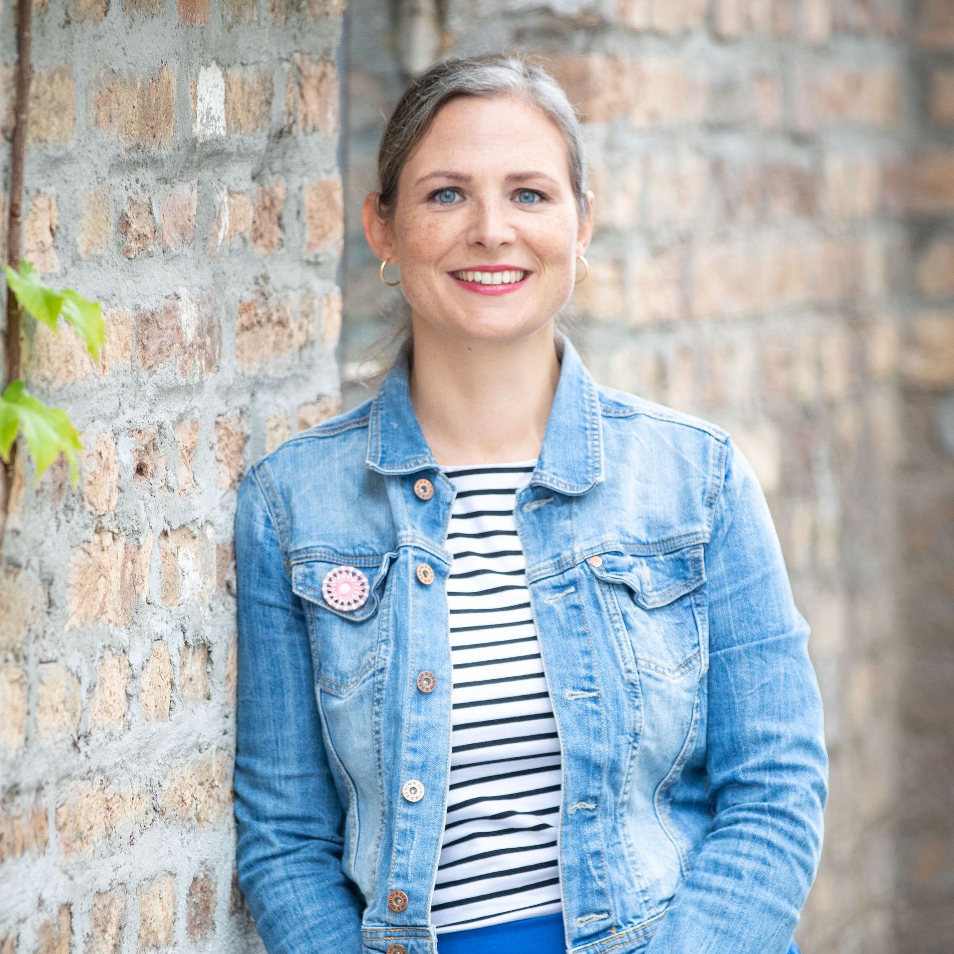 Frau in Jeansjacke, gestreiftem Hemd und blauem Rock lächelt in die Kamera und lehnt an einer Backsteinmauer.