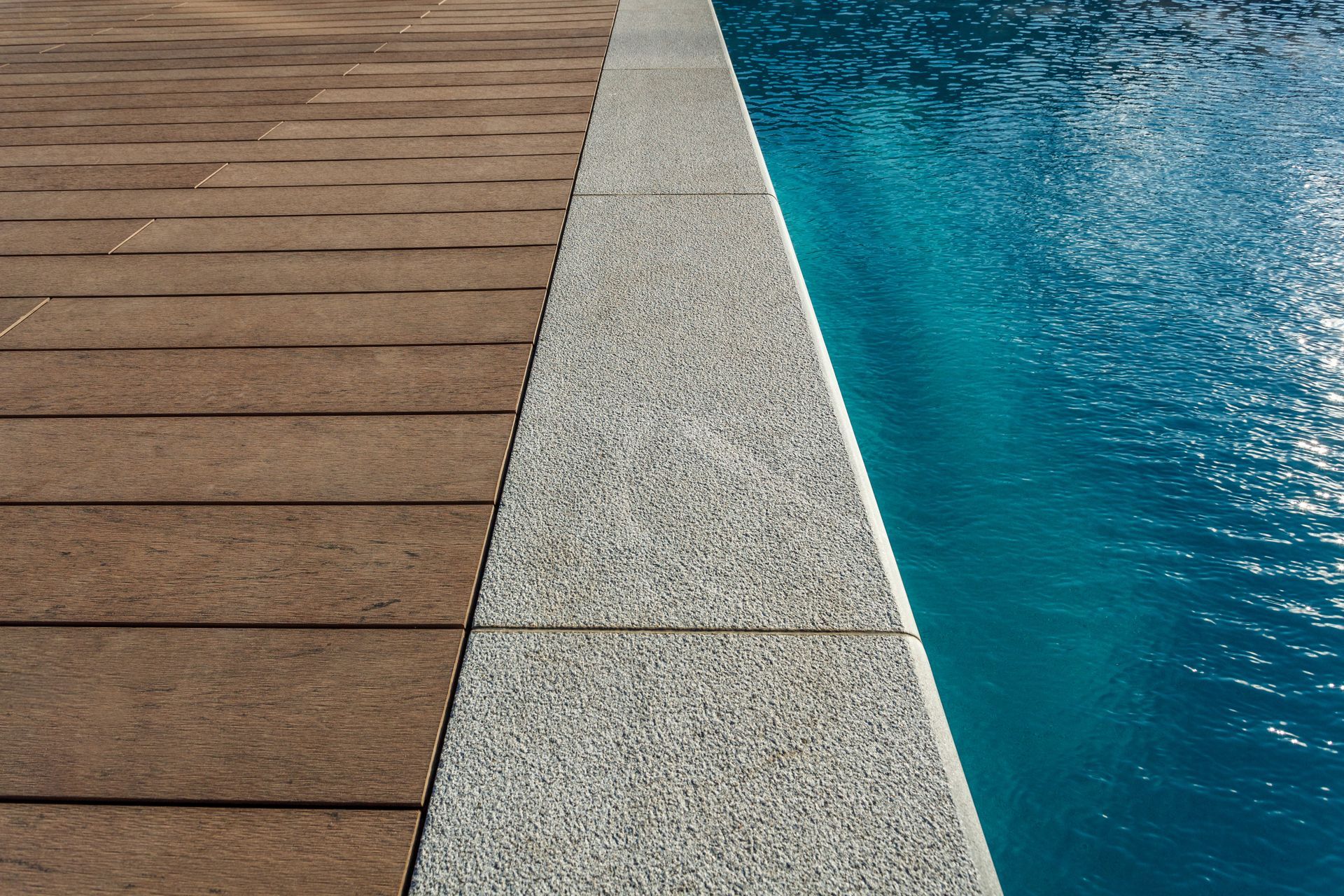 Poolside view with brown paving stones, speckled concrete border, and turquoise water.