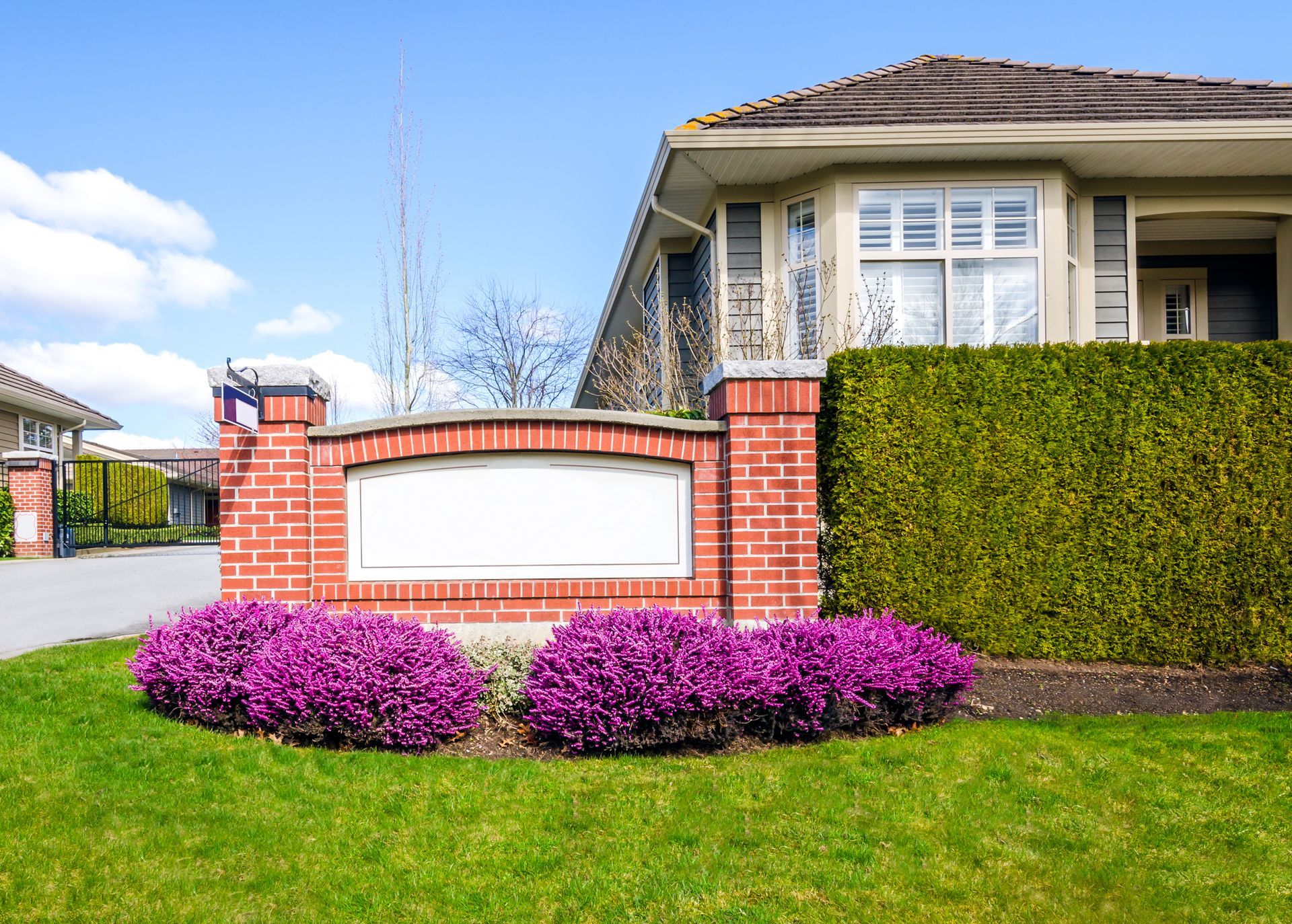 Brick entrance sign with blank space, flanked by pink flowers and green hedge in front of a house.