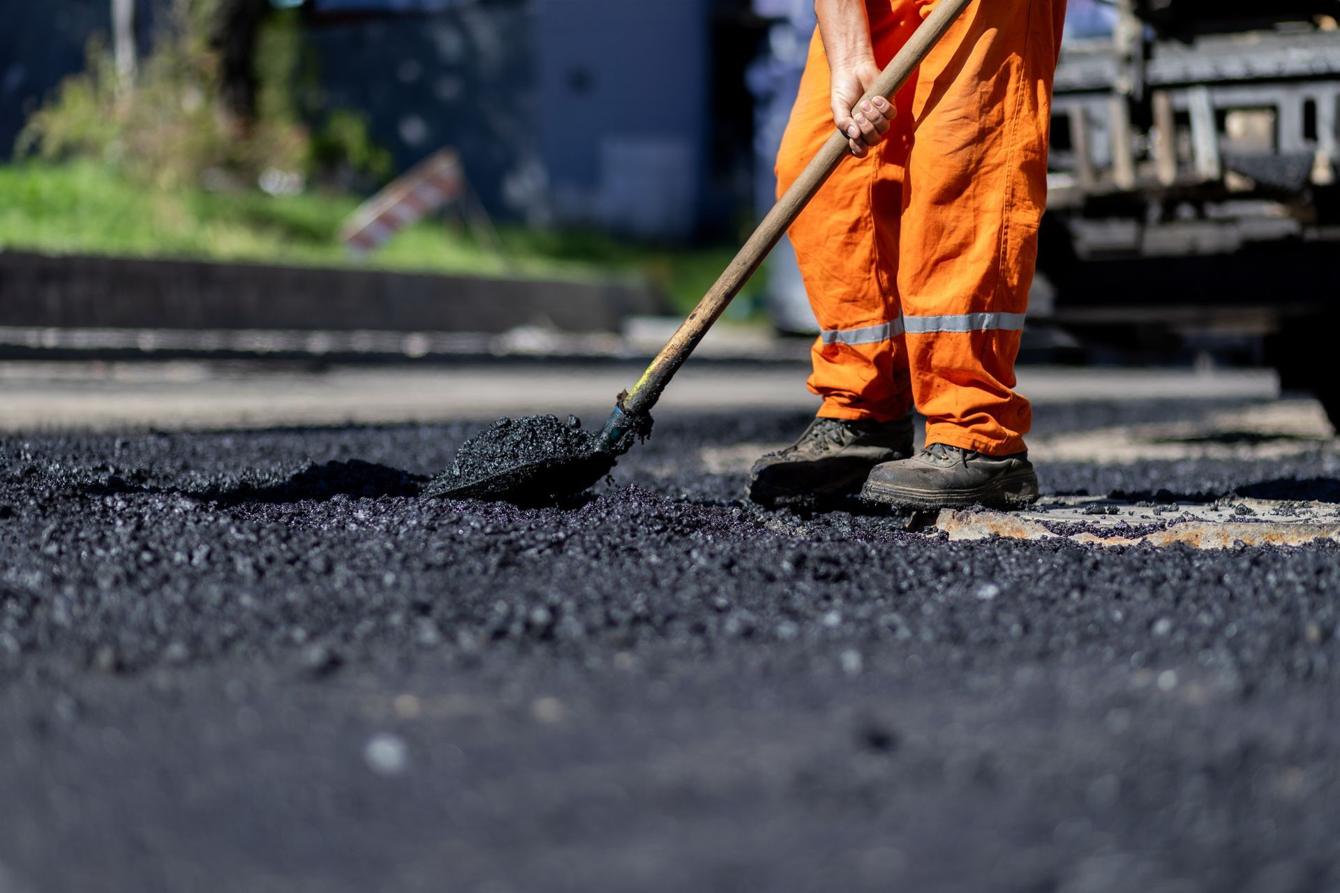 Construction worker in orange overalls raking hot asphalt on a road.