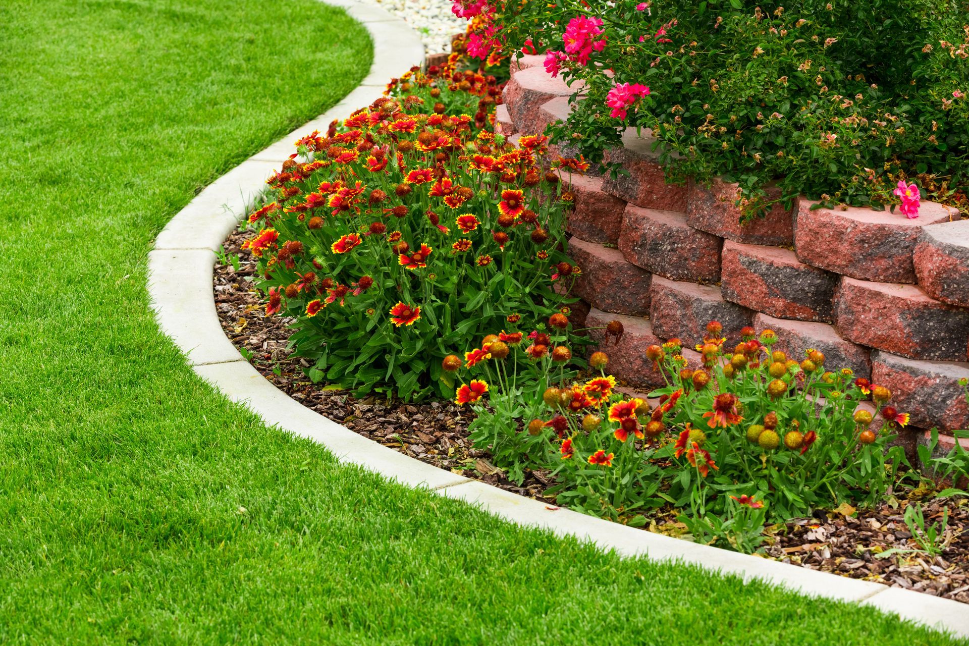 Lush green lawn borders a curved flower bed with red and yellow flowers and a stacked brick wall.