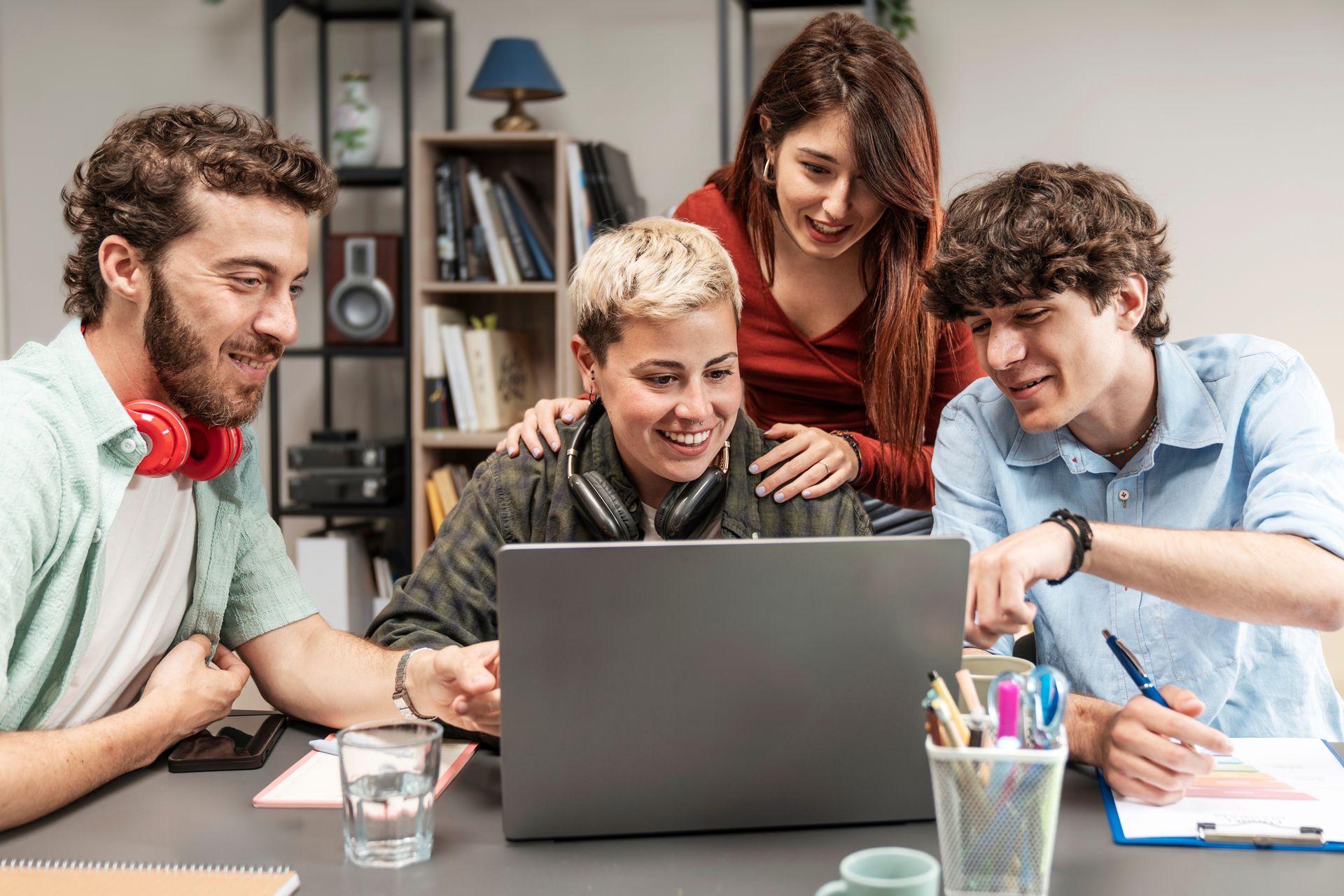Four young adults collaborating around a laptop, smiling and pointing, in a casual office setting.