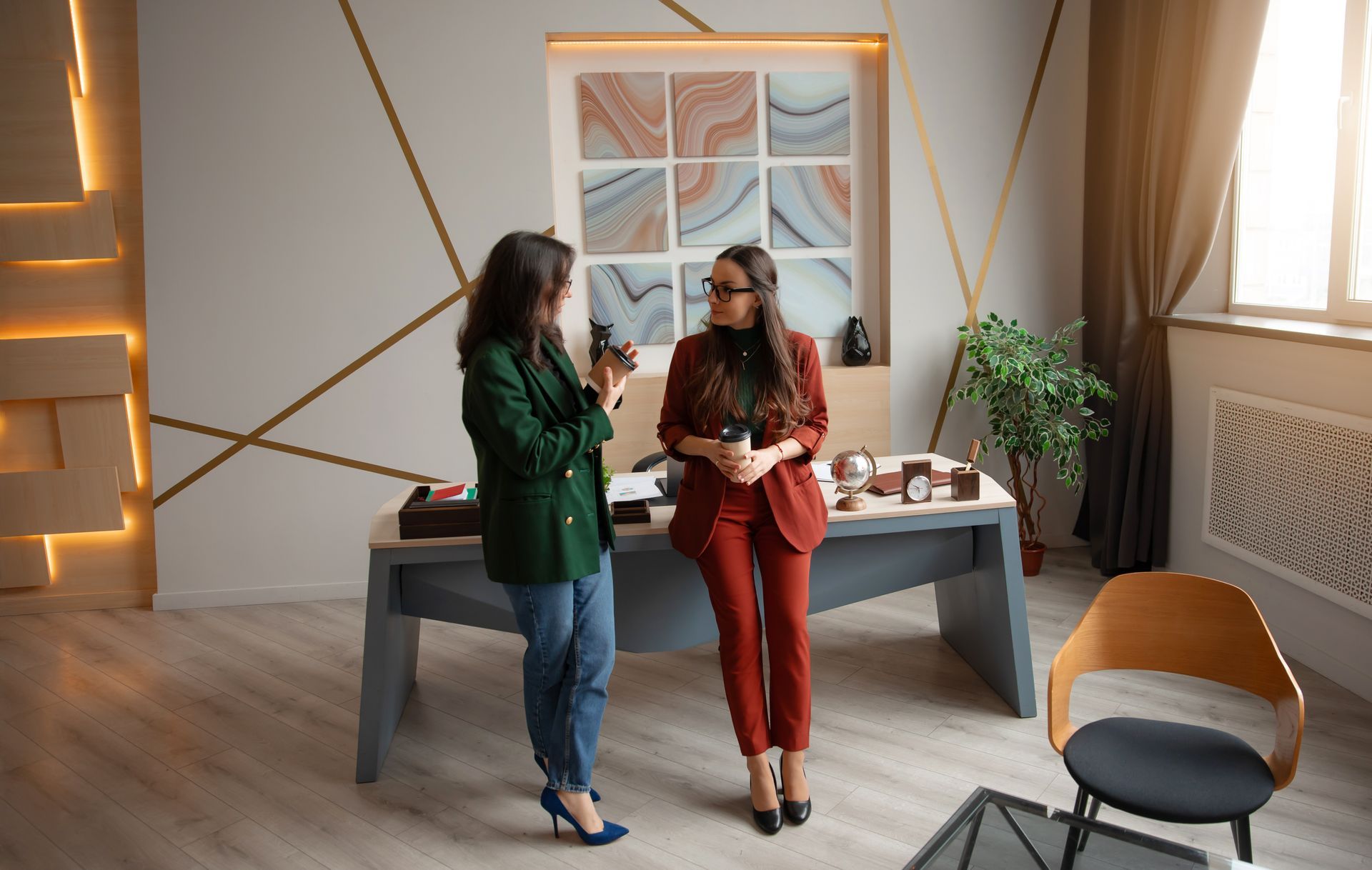 Two women in a stylish office; one in red suit holding a coffee, the other in green jacket looking at a phone.
