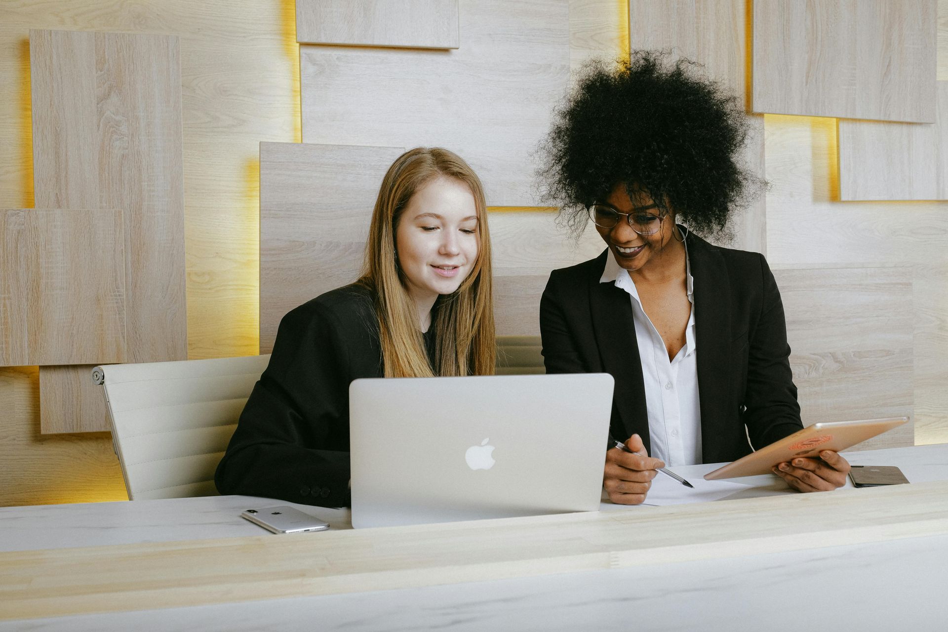 Two women in business attire, looking at a laptop, smiling in a wood-paneled office.