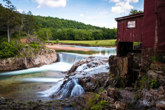 Waterfall flowing beside a red mill by a forested riverbank under a blue sky