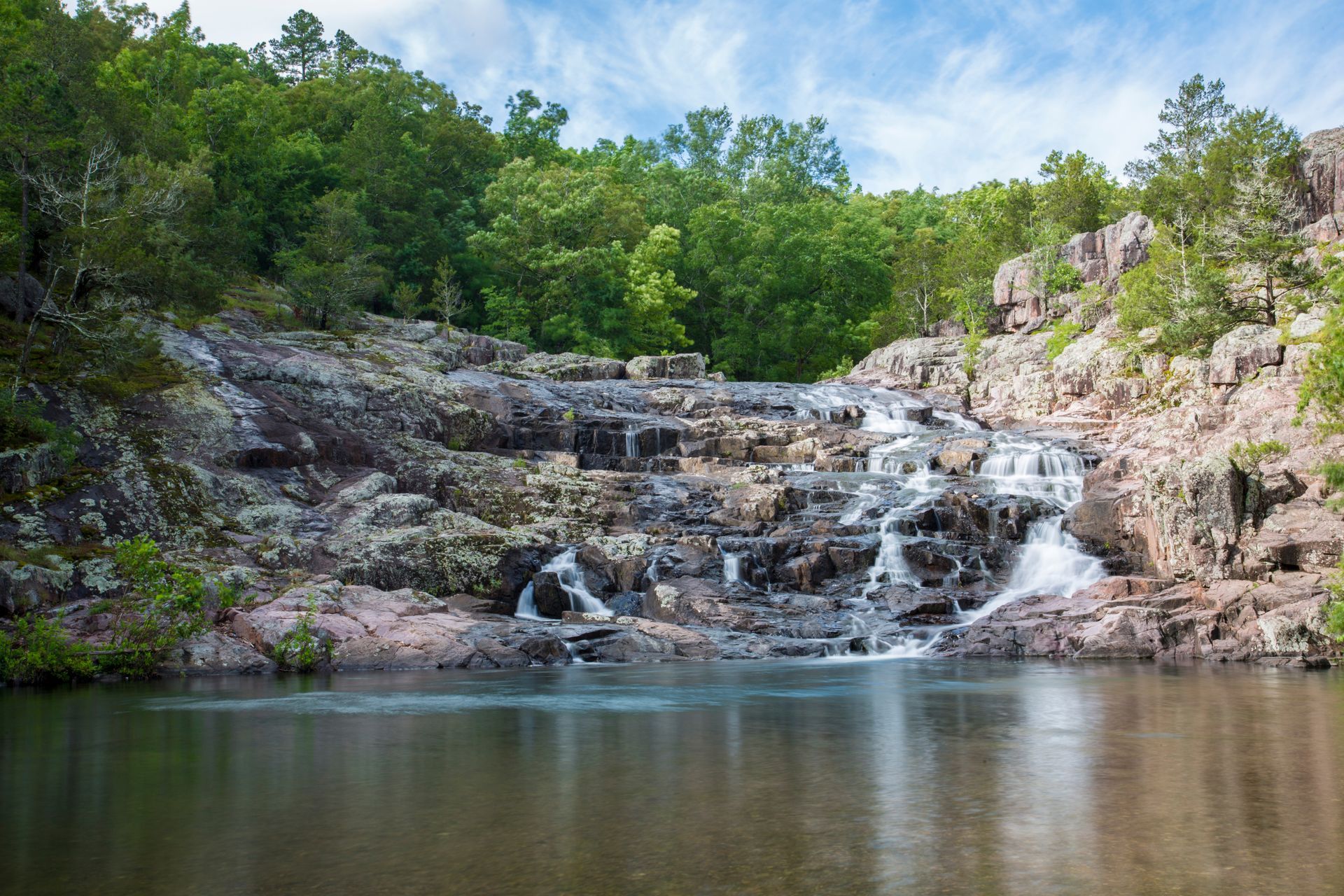 Rocky waterfall cascading into a calm pool, surrounded by lush green trees under a blue sky.