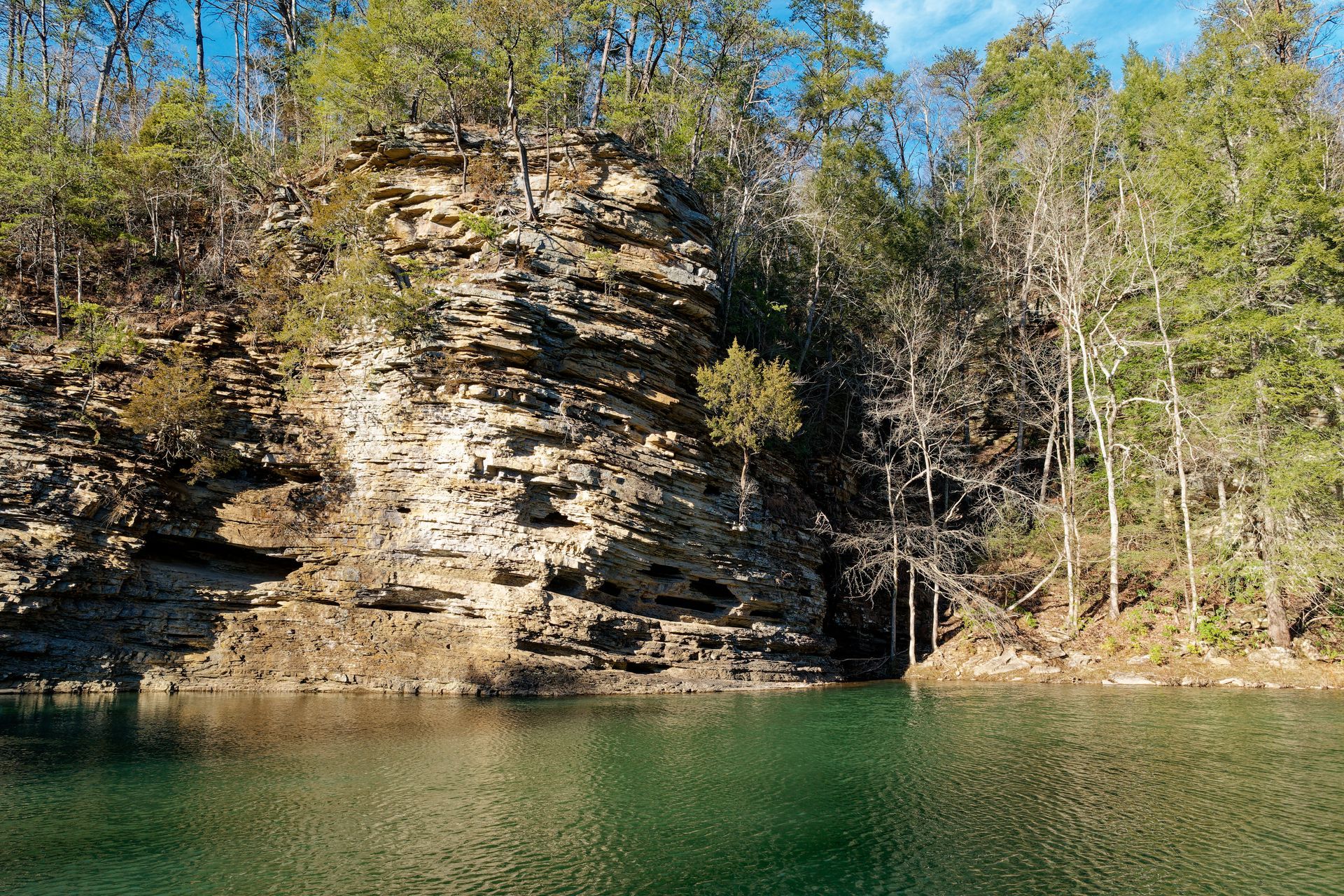 Rocky cliff above calm green water, with trees in autumn foliage along the shoreline