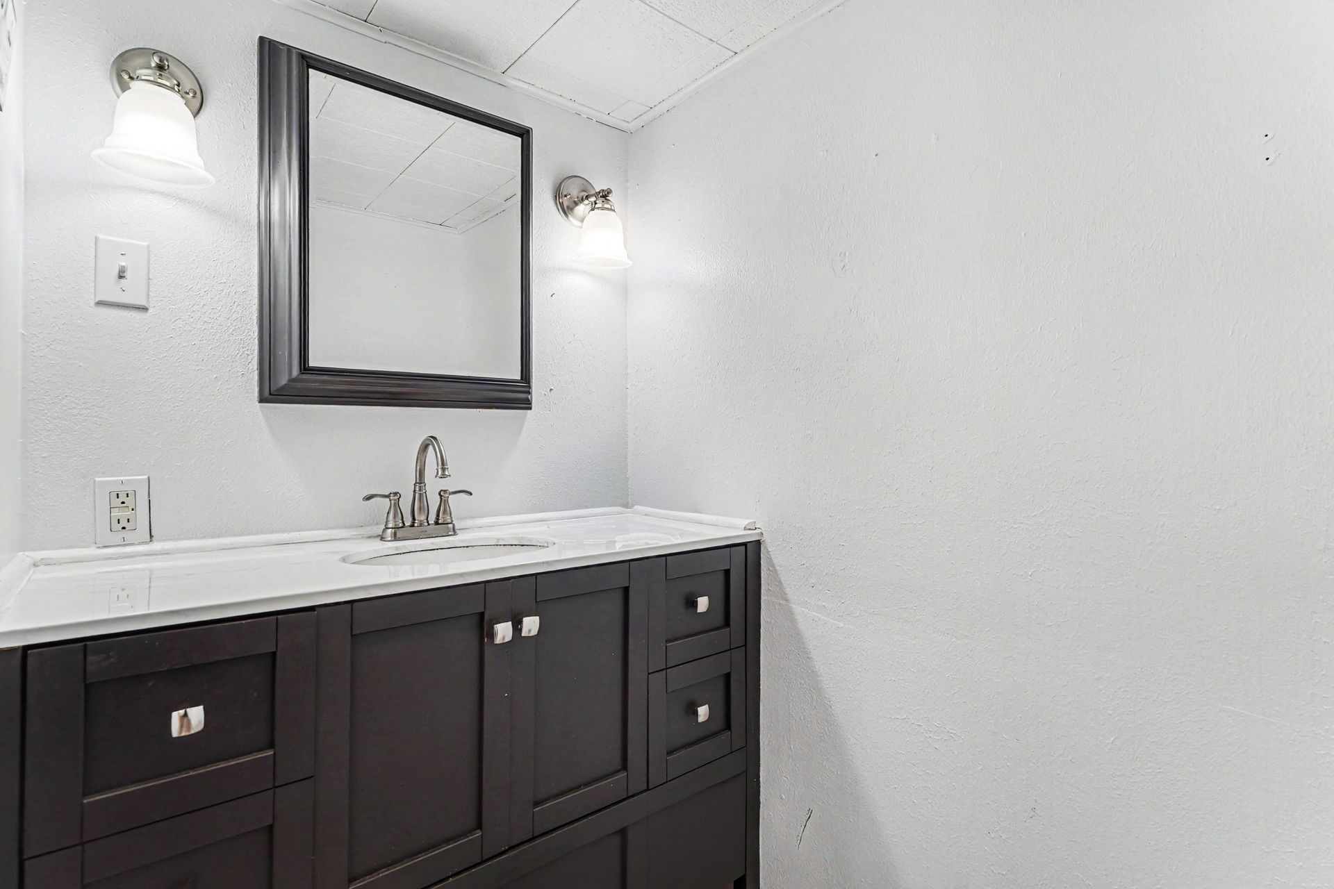 Bathroom vanity with dark cabinets, white countertop, mirror, and wall sconces against a textured white wall