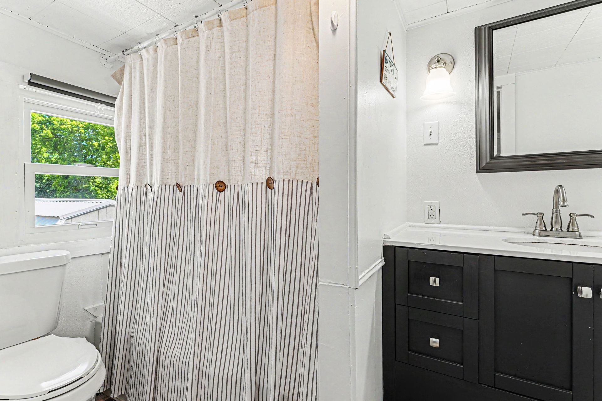 White bathroom with shower curtain, toilet, black vanity, sink, and window with green view