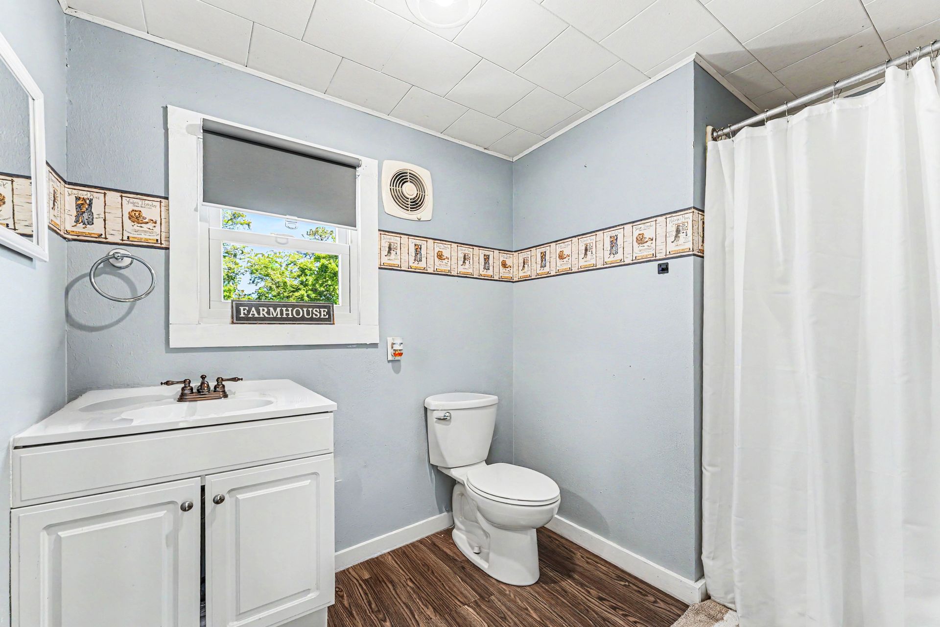 Small bathroom with white vanity, toilet, and shower curtain; light blue walls and wood-look floor.