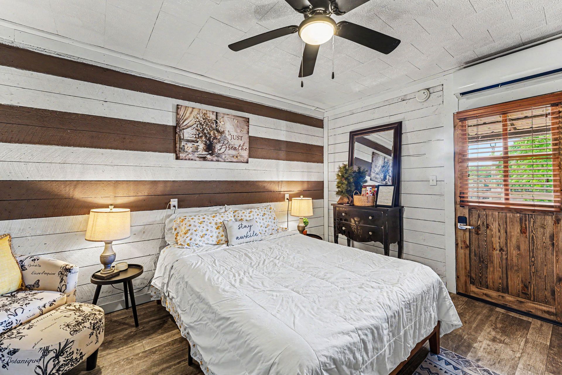Rustic bedroom with white bedspread, wood accent wall, ceiling fan, lamp, mirror, and barn-style door