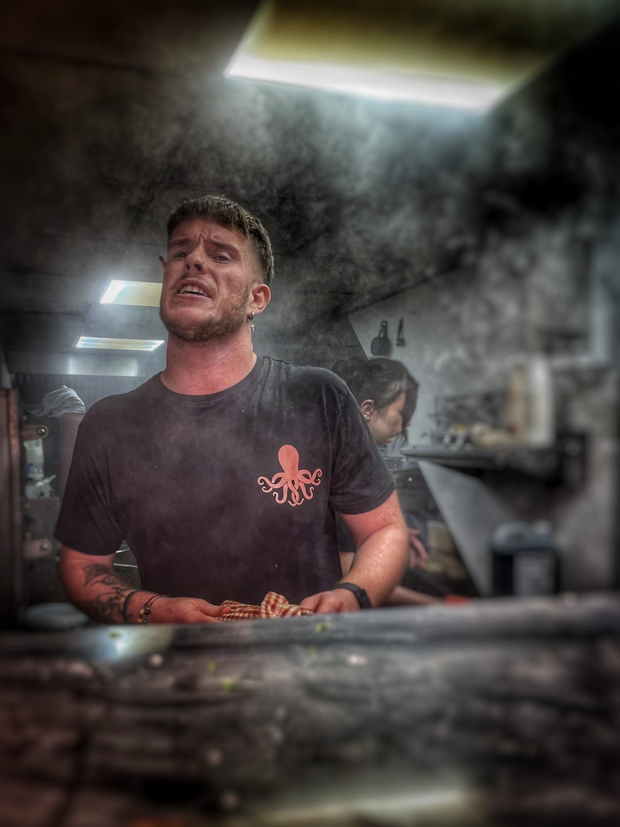 A man wearing a black shirt with an octopus on it is cooking food in a kitchen.