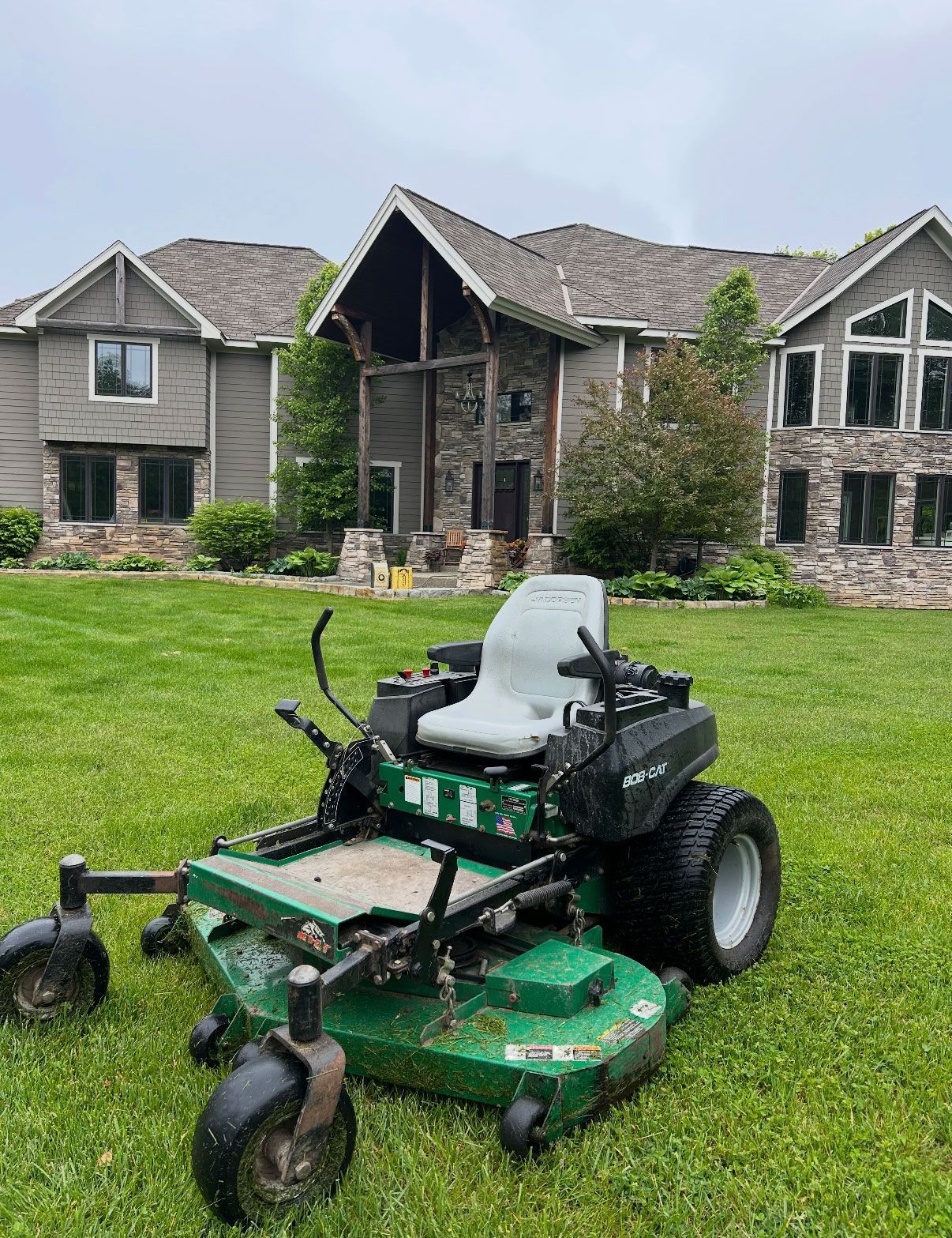 Green riding lawnmower on a lawn in front of a large house. Overcast sky.