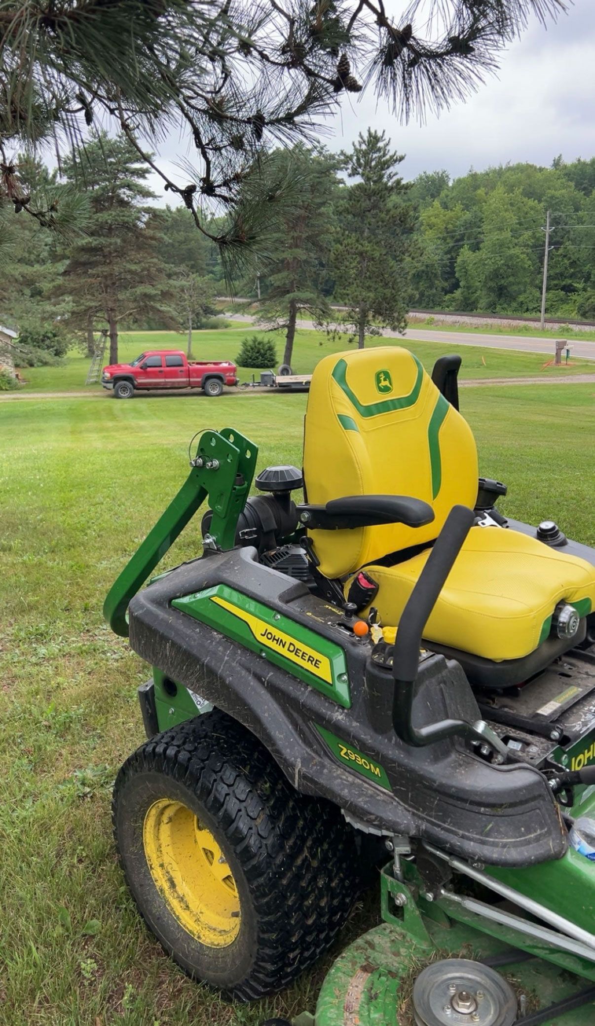 John Deere zero-turn lawnmower on grass, trees, and red truck in background. Yellow and green colors.