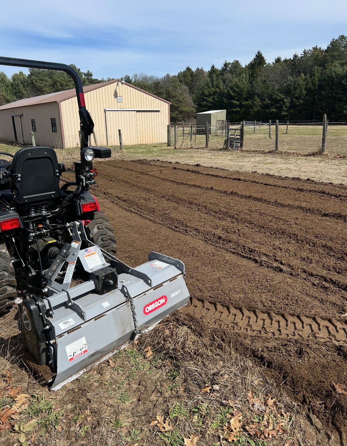 A tractor with a tiller working soil in a field near a barn on a sunny day.