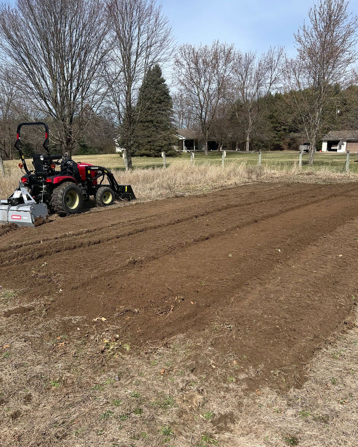 Red tractor tilling a field of brown soil in a yard. Trees and a small building in the background.