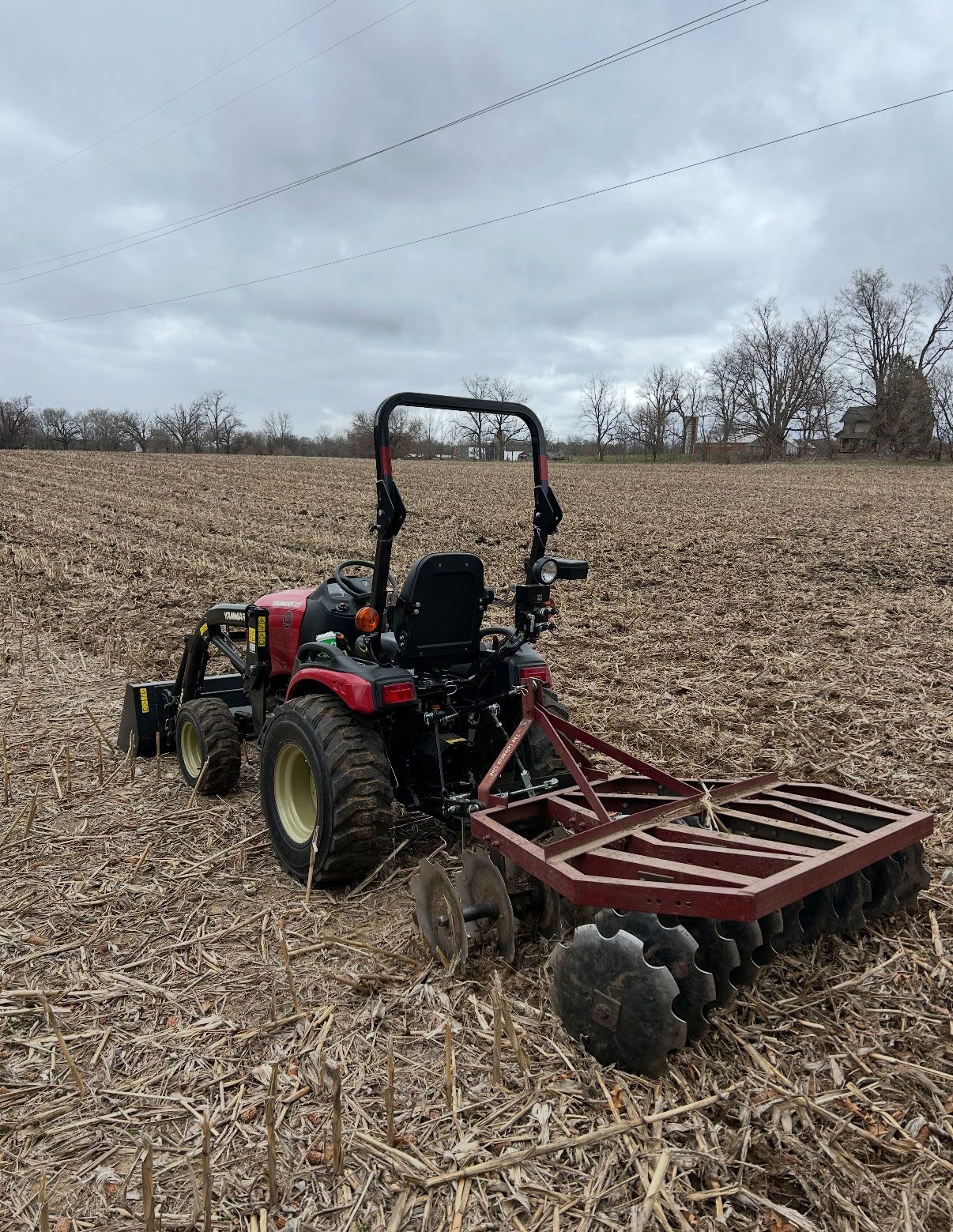 A red tractor with disc harrow plowing a field under a cloudy sky.