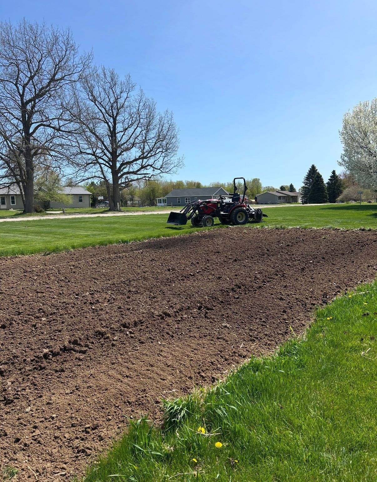 Tilled garden bed with a tractor in a grassy yard under a bright blue sky.
