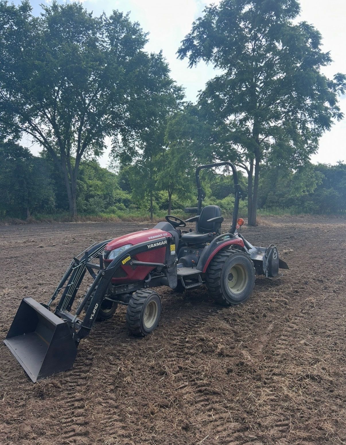 Red tractor with front loader and rear tiller on a plowed field in front of a tree line.