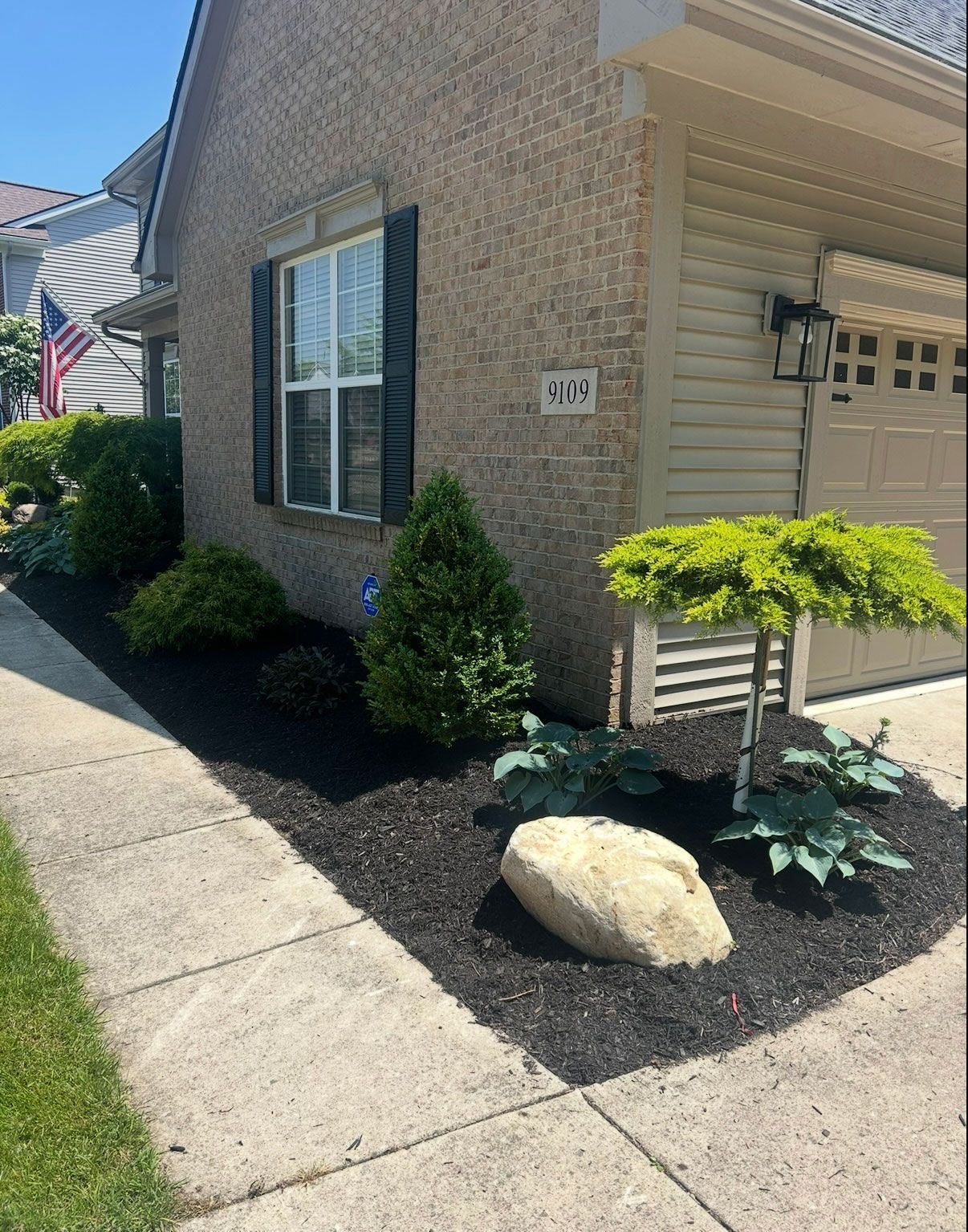 Landscaped yard with black mulch, shrubs, a large rock, and a tree next to a house with a garage.