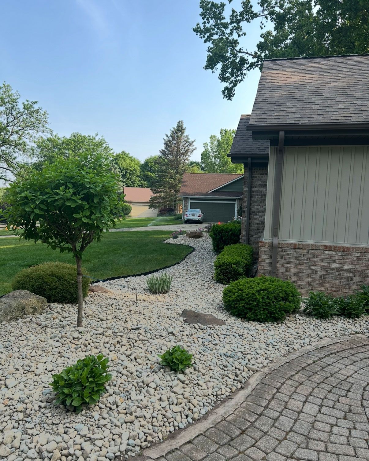Landscaped front yard with a stone path, small tree, and shrubs next to a house with a green roof.