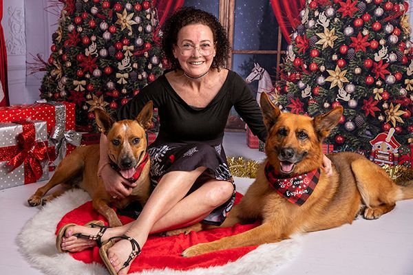 A woman is sitting on the floor with two dogs in front of a christmas tree.