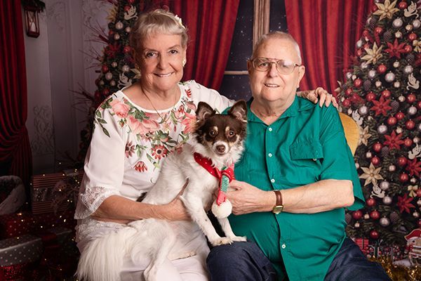 A man and woman are posing for a picture with their dog in front of a christmas tree.